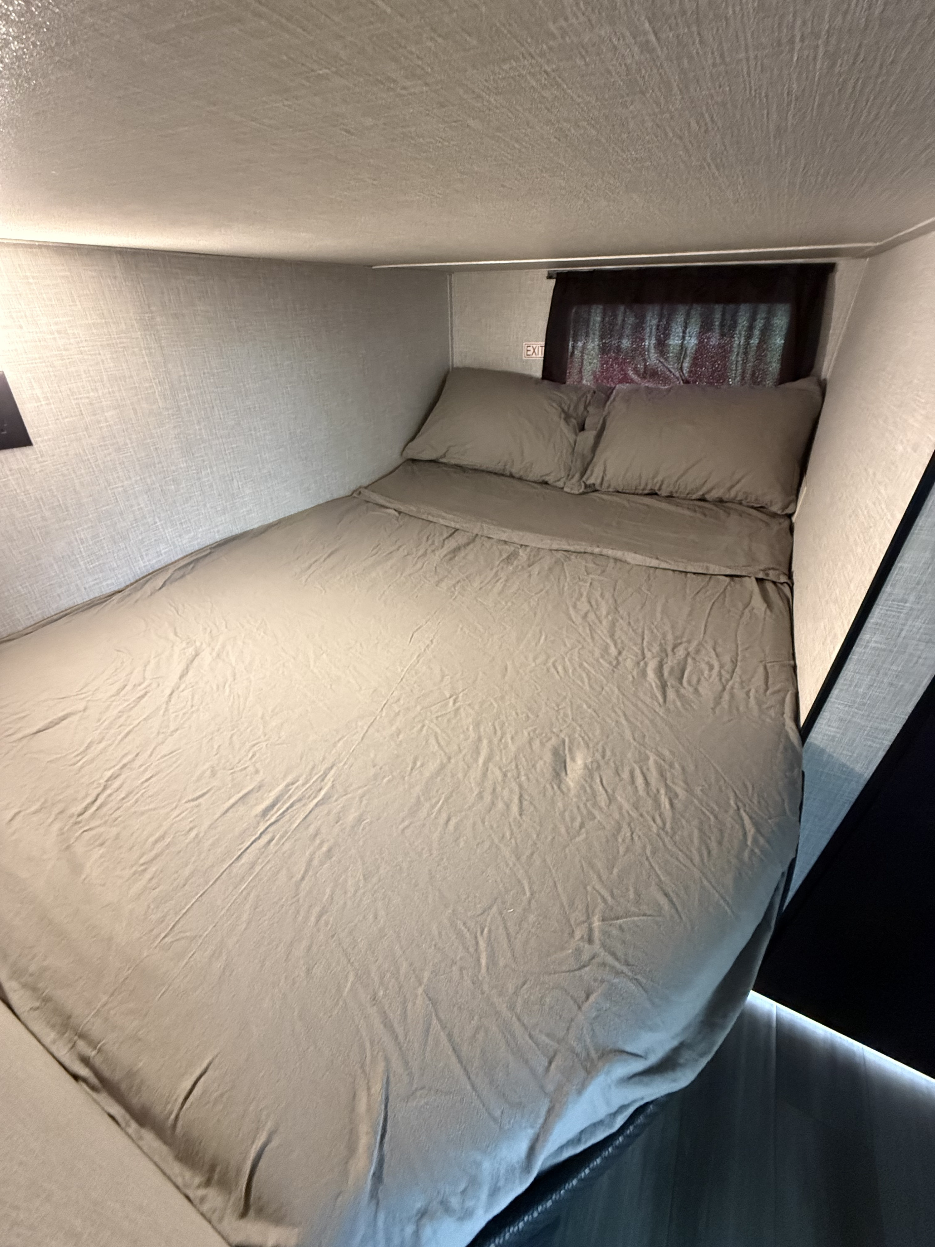 Cozy gray bed with pillows in an RV rental sleeping area, featuring an exit sign above the headboard and a window.. Jayco Jay Flight SLX 2024