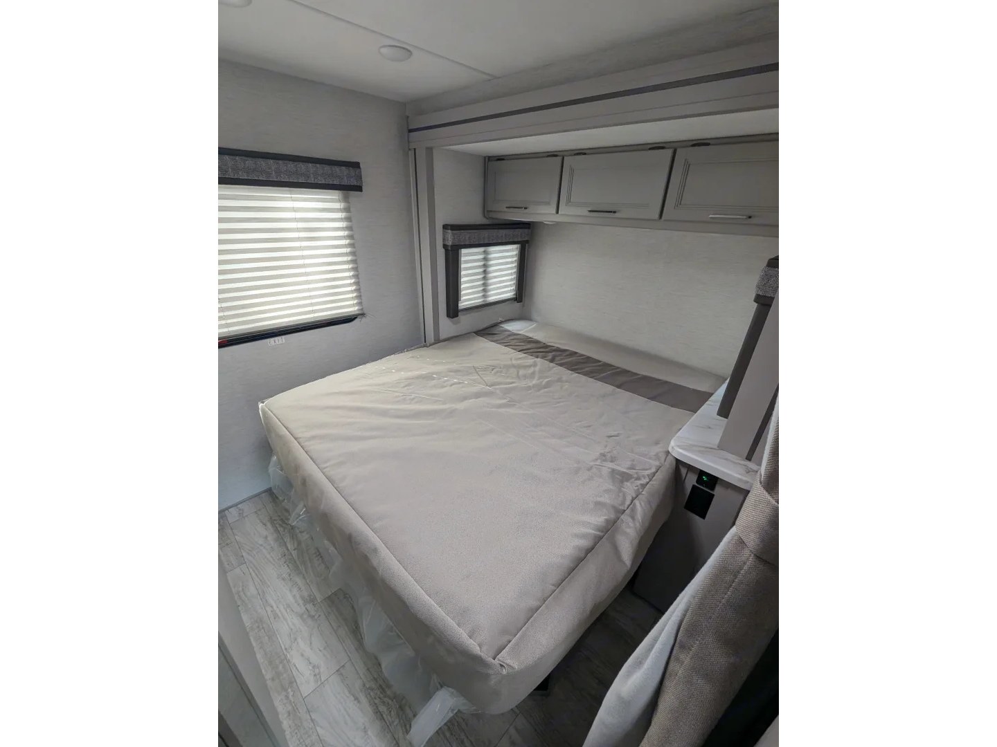 RV rental bedroom interior with a light-colored bed under protective wrap, overhead cabinets, windows with blinds, and power outlets.. Thor Chateau 2025