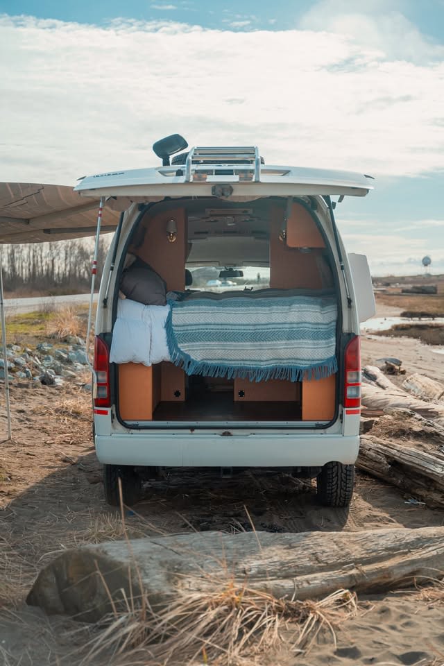 Discover adventure with this rv rental campervan! Cozy bed visible through the open tailgate, parked on a sandy beach.. Toyota Bus 4x4 Campervan conversion of Toyota Hiace 2007
