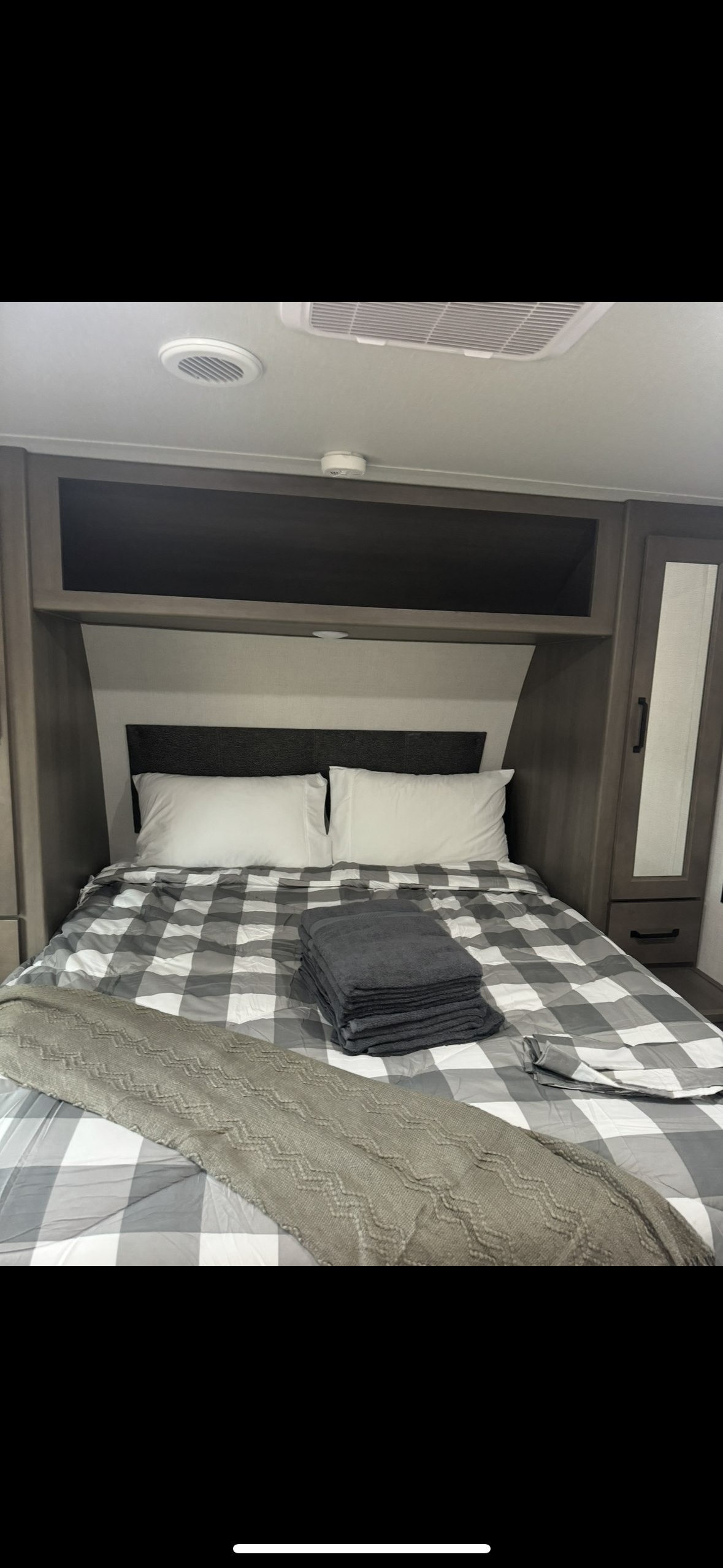 Cozy RV rental bedroom features checkered bedding, stacked towels, and practical overhead storage for comfortable travel.. Grand Design Transcend Xplor 2024