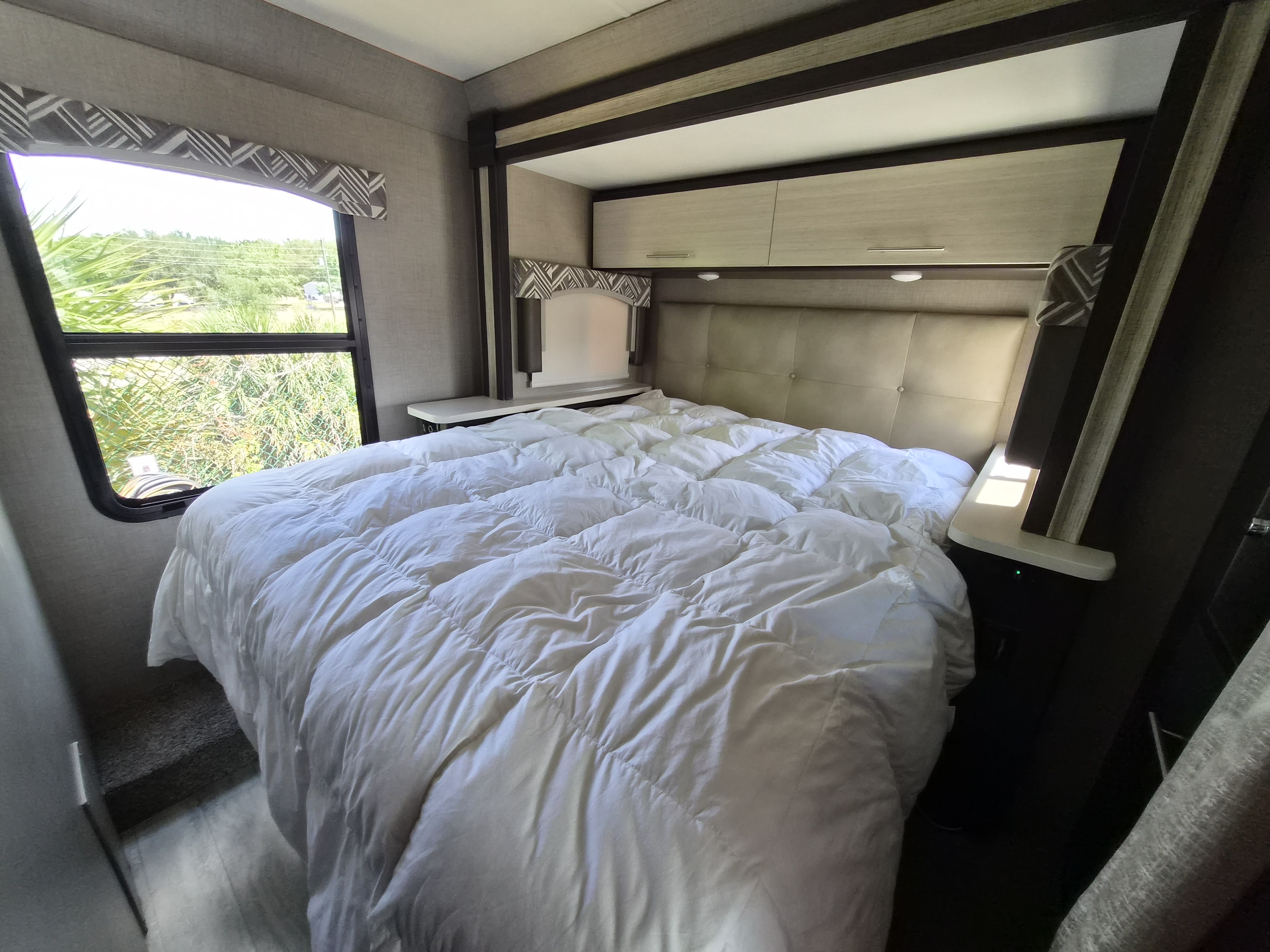 Inviting RV rental bedroom with a large white comforter, functional overhead cabinets, and a window showcasing outdoor greenery.. Thor Axis 2020