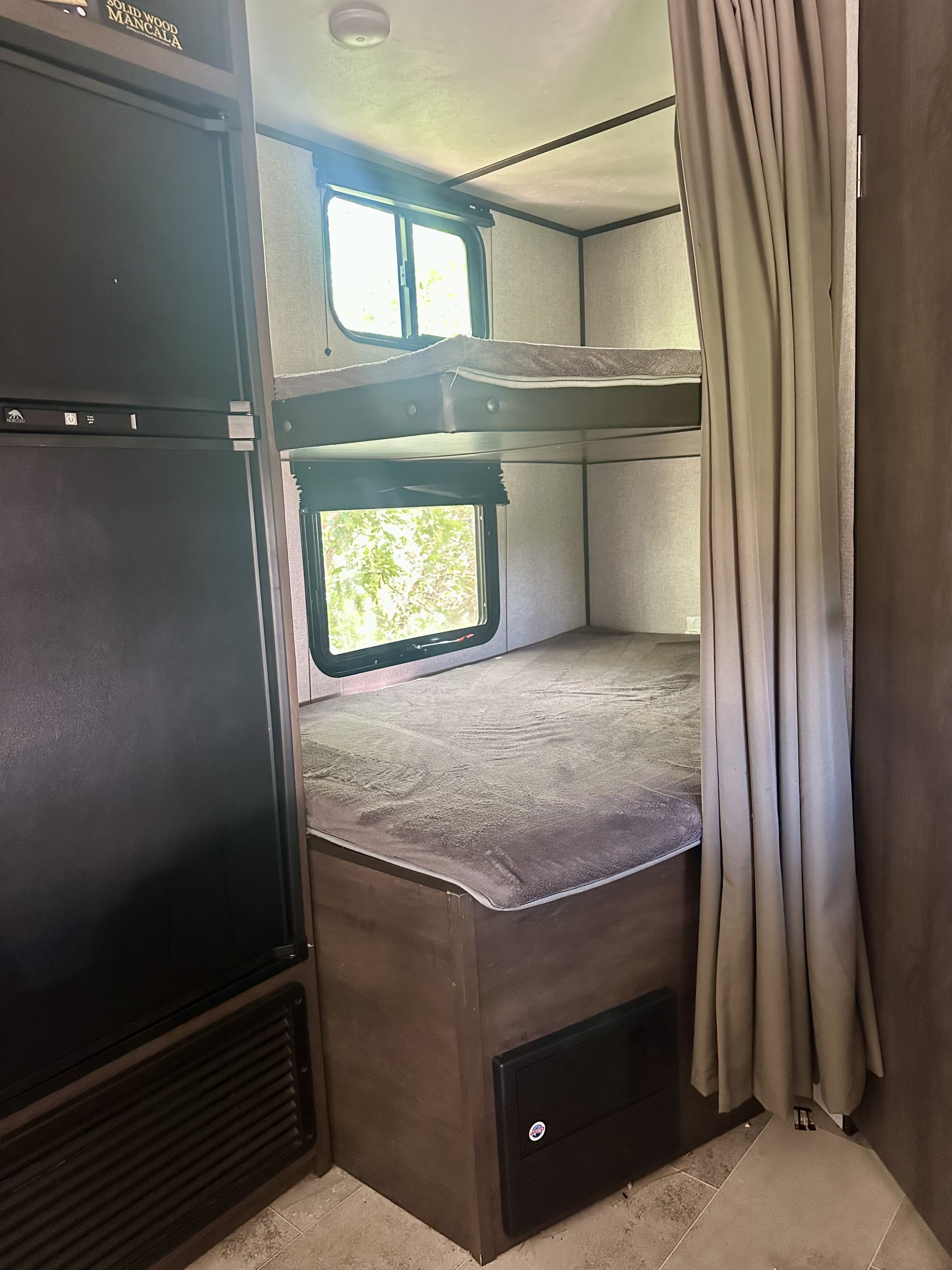 Inside an RV rental: two comfy bunk beds with windows, next to a refrigerator and a privacy curtain.. Dutchmen Coleman Lantern 2020