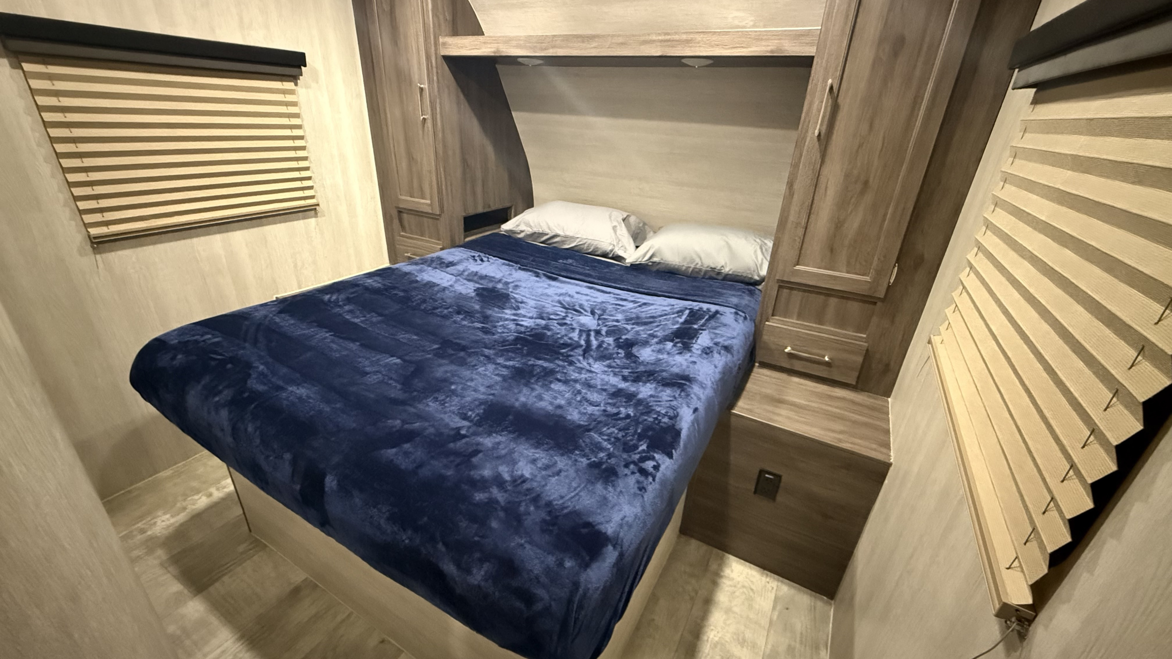 Cozy RV rental bedroom featuring a bed with a blue blanket, wood cabinetry, and window blinds. A compact, functional interior.. Genesis Supreme Rv Genesis Supreme 2022