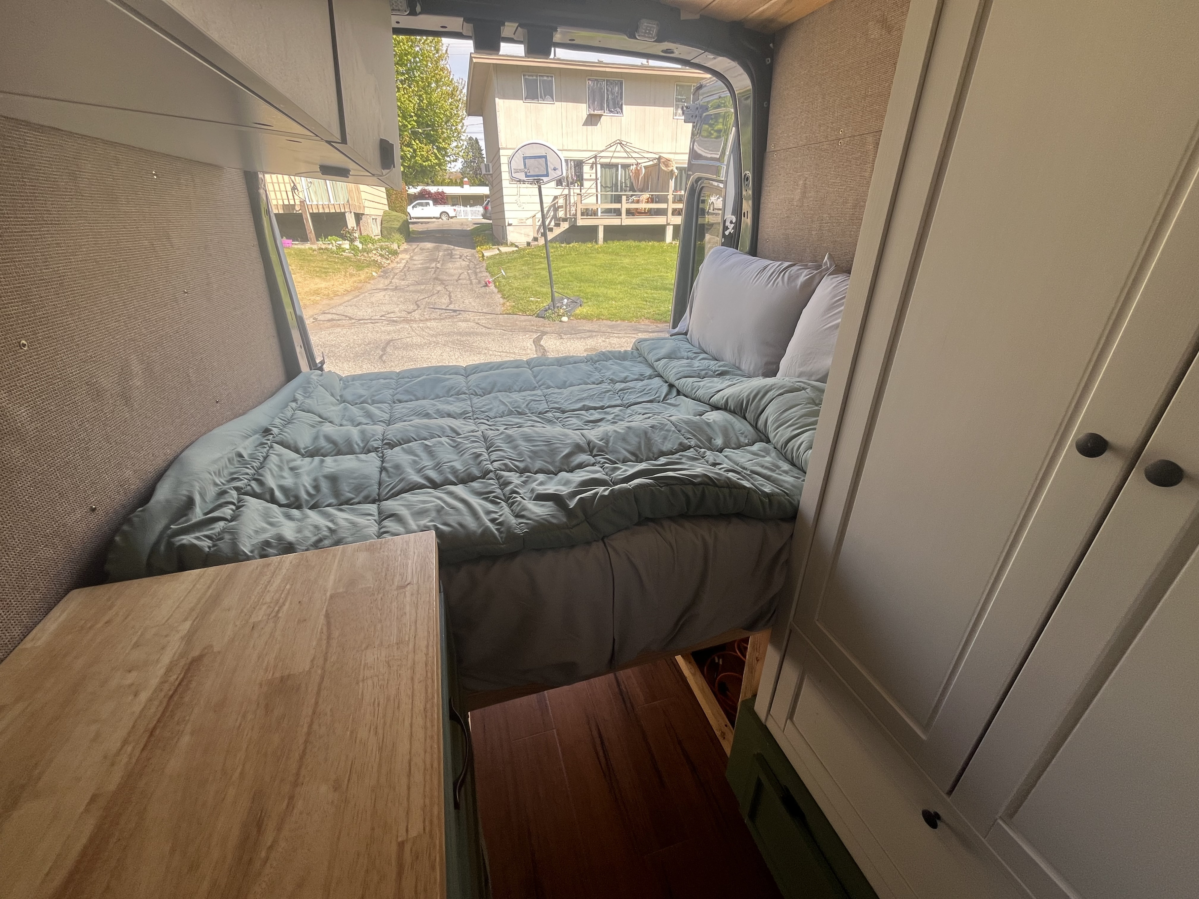 RV rental van interior: comfortable bed, pillows, wooden cabinetry, and open rear doors revealing a residential driveway view.. Ford Custom Transit 2020