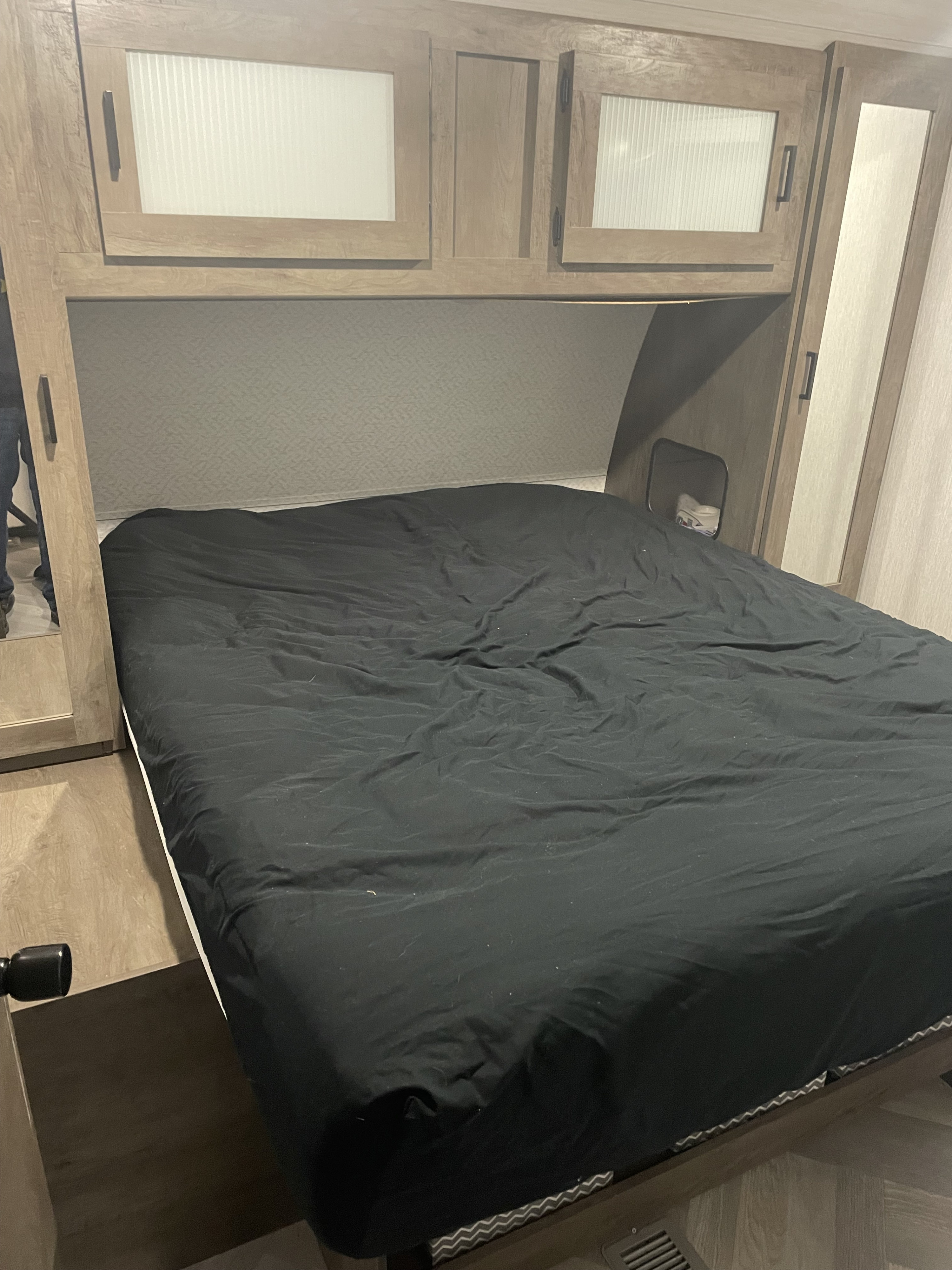 An RV rental's bedroom shows a bed with dark bedding, overhead wooden cabinets, and a side storage cubby. Modern camper interior.. Forest River Salem 2022