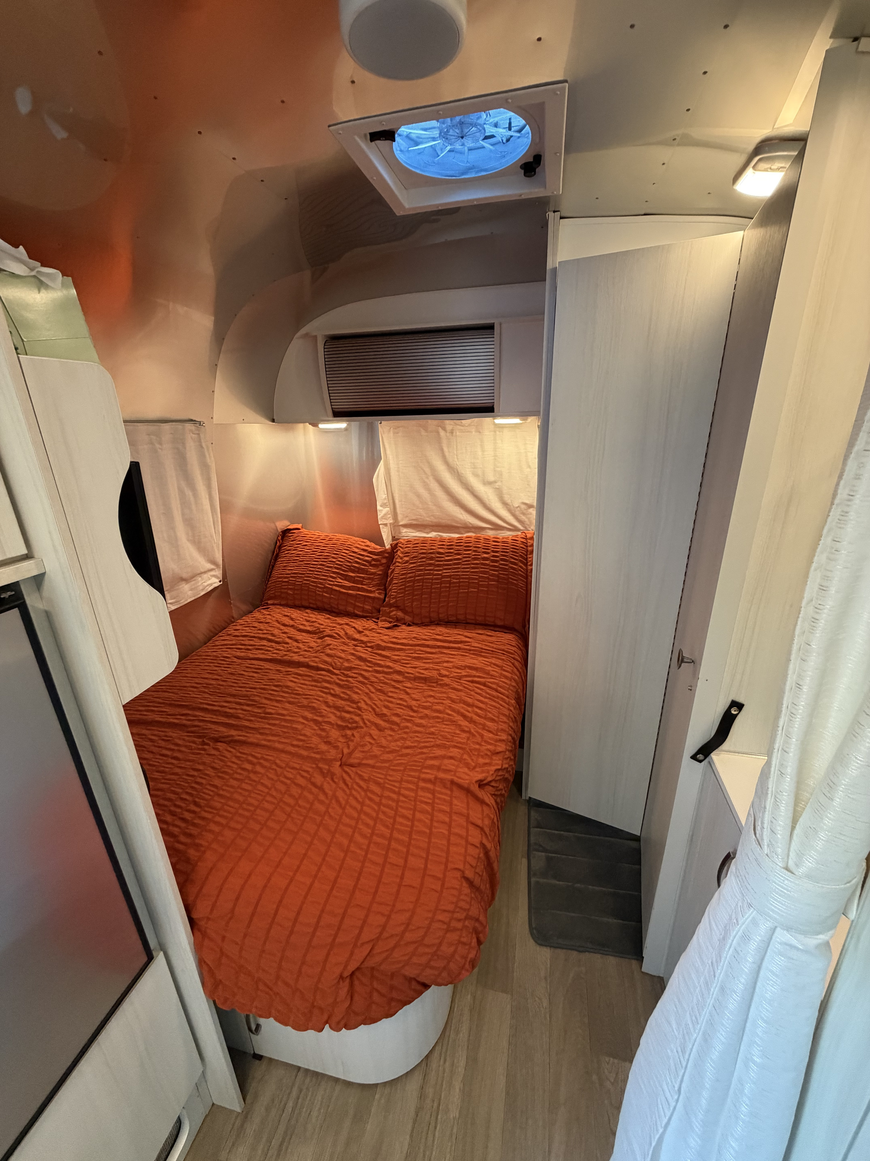 This compact rv rental bedroom features vibrant orange bedding, polished metallic walls, and a ceiling vent, offering cozy comfort.. Airstream Bambi 2021