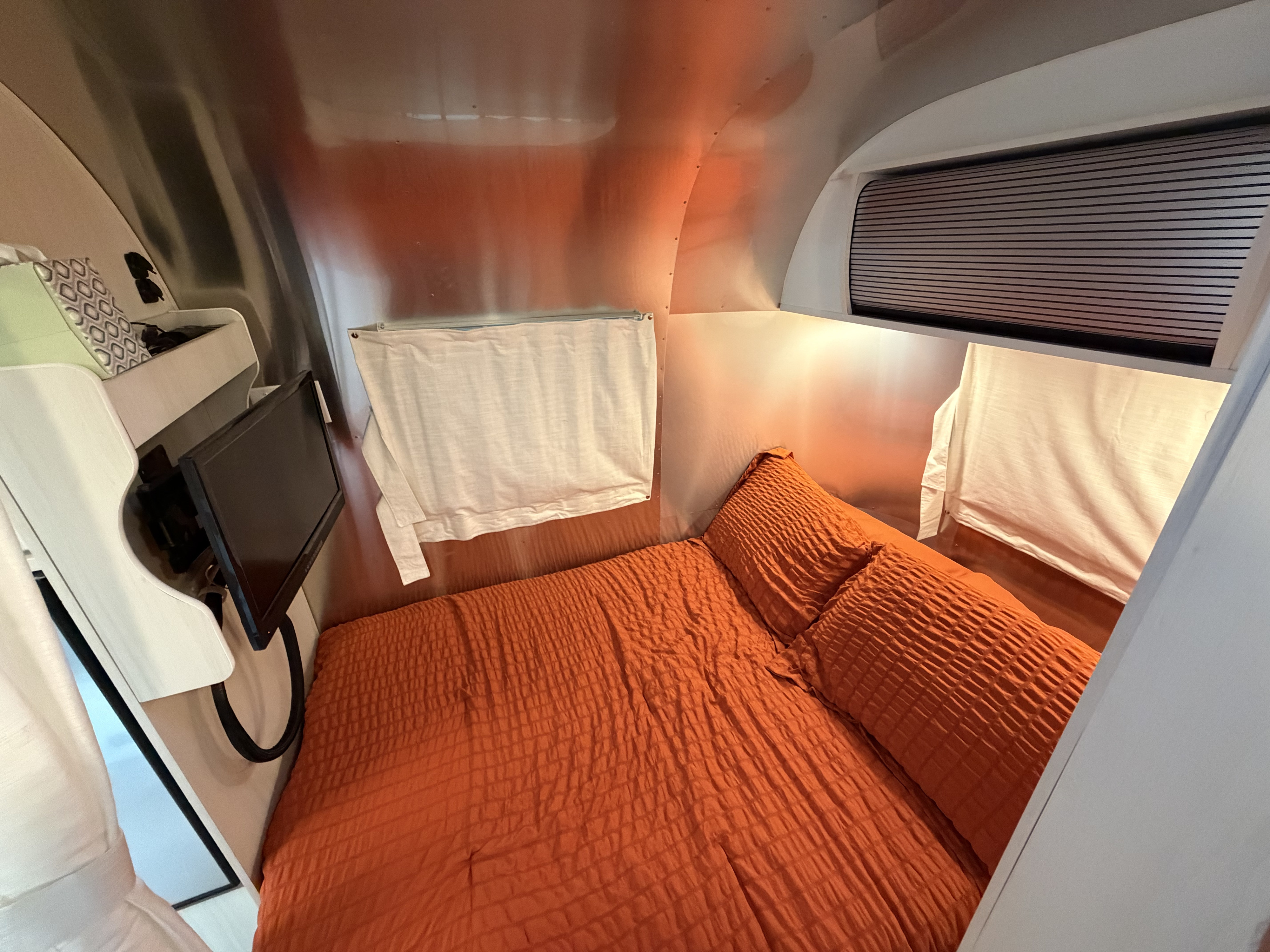Cozy RV rental bedroom featuring an orange bed, small TV, and shiny metallic walls for a modern travel experience.. Airstream Bambi 2021