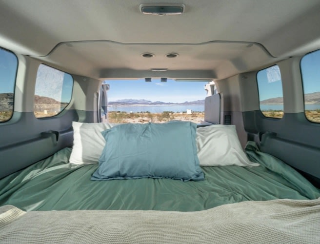 Interior of an rv rental van: a comfortable bed with green sheets & pillows, facing a scenic lake and mountains.. Ford Transit 2017