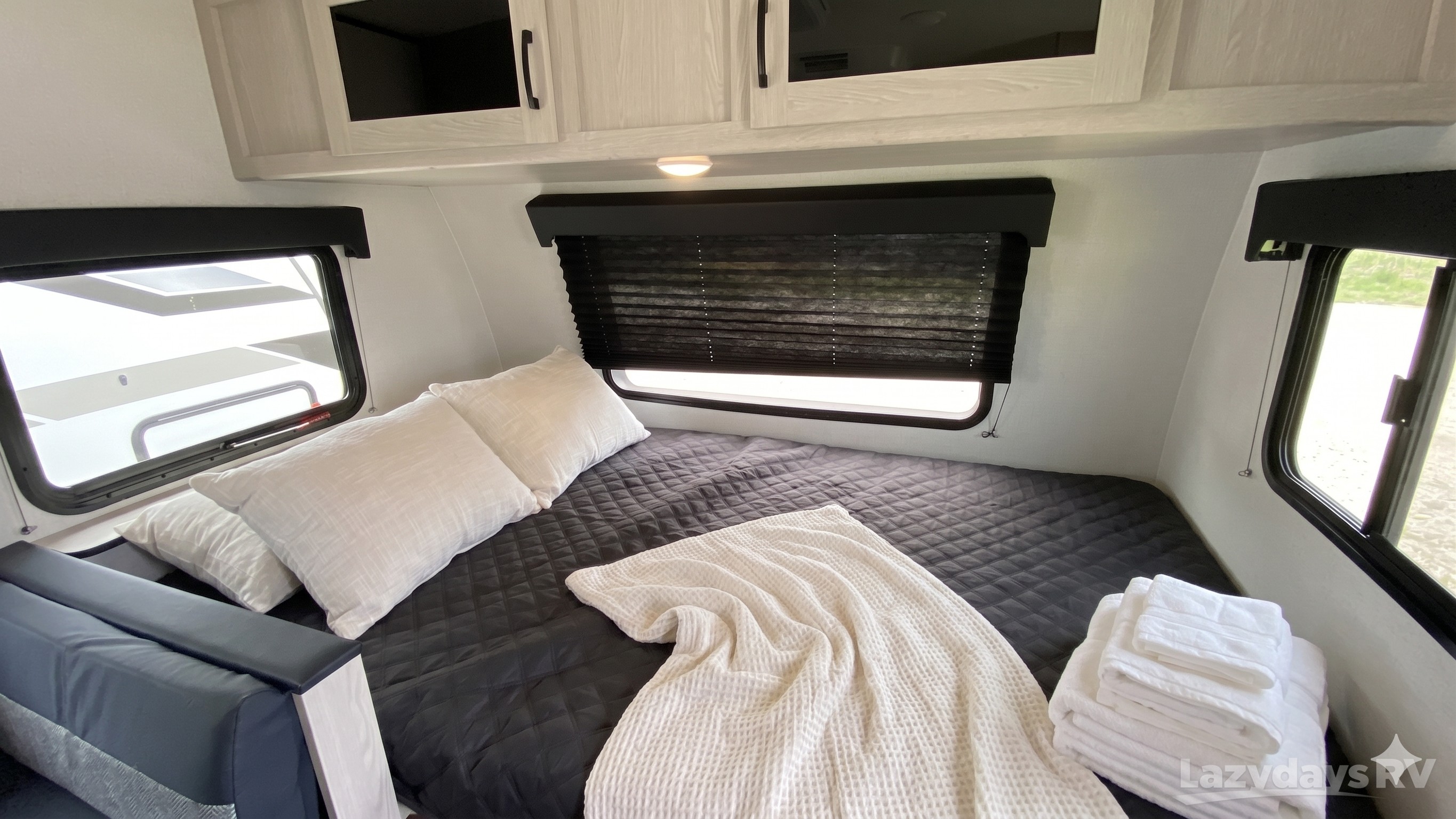 A bright, cozy RV rental bedroom features a bed with dark quilt, white linens, overhead cabinets, & windows.. Coachmen Apex Nano 2023