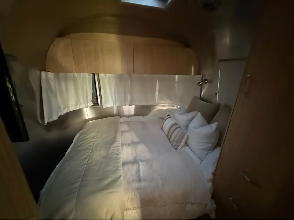 Cozy and bright bedroom inside an RV rental, featuring a comfortable bed with white linens, pillows, and wooden accents.. Airstream Flying Cloud 2018