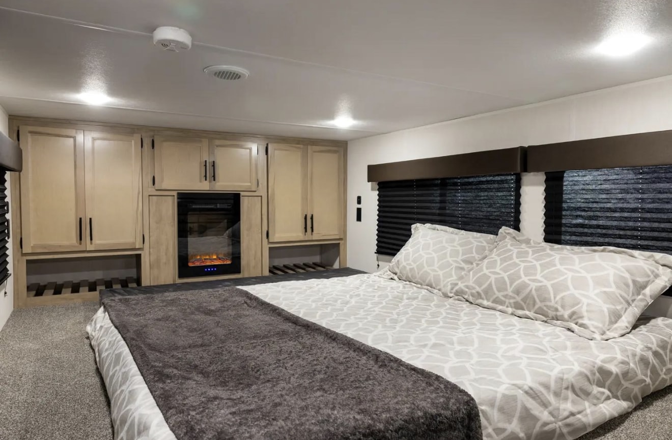 Modern RV rental bedroom with a large bed, light wood cabinets, electric fireplace, and dark window blinds for comfortable travel.. Forest River Cherokee Timberwolf 2025