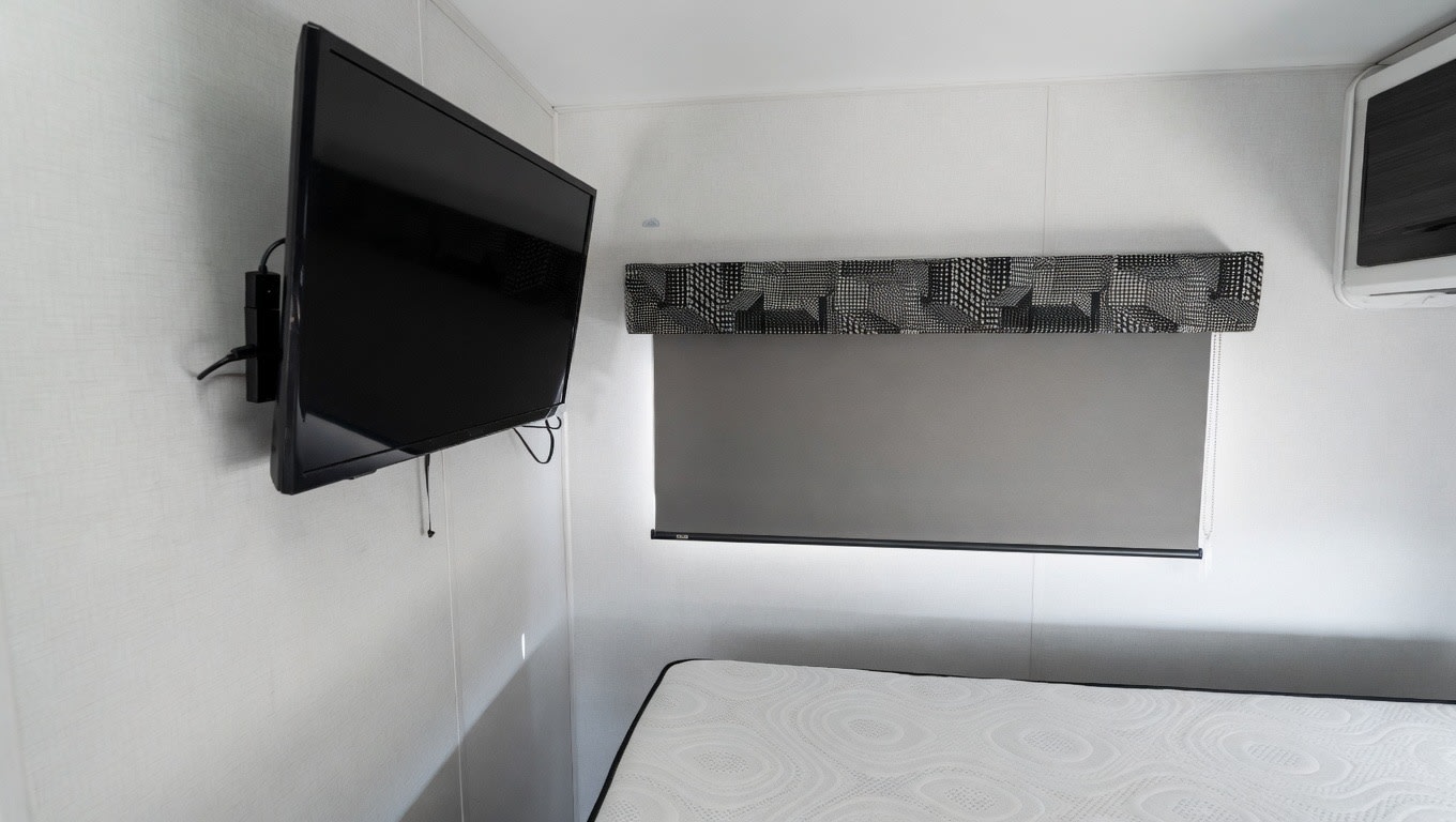 RV rental bedroom interior featuring a wall-mounted TV, window with patterned valance and grey shade, and a comfortable mattress.. Forest River Sunseeker LE 2023