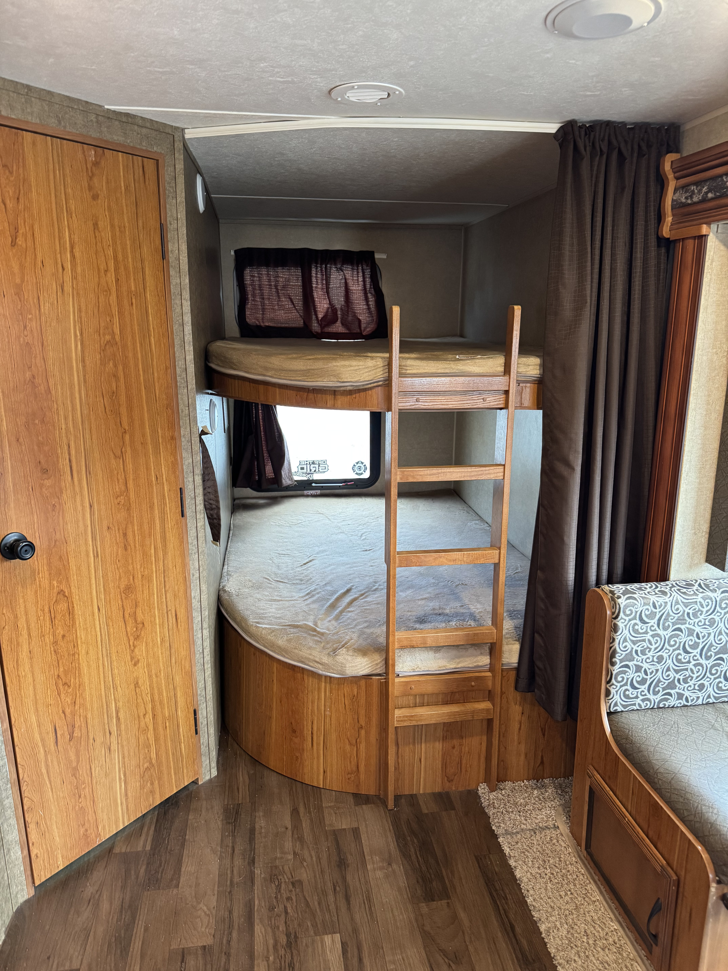 RV rental interior with stacked bunk beds, a wooden ladder, and a door on the left. Part of a patterned sofa is on the right. Wooden walls & floor.. Starcraft Launch 2016