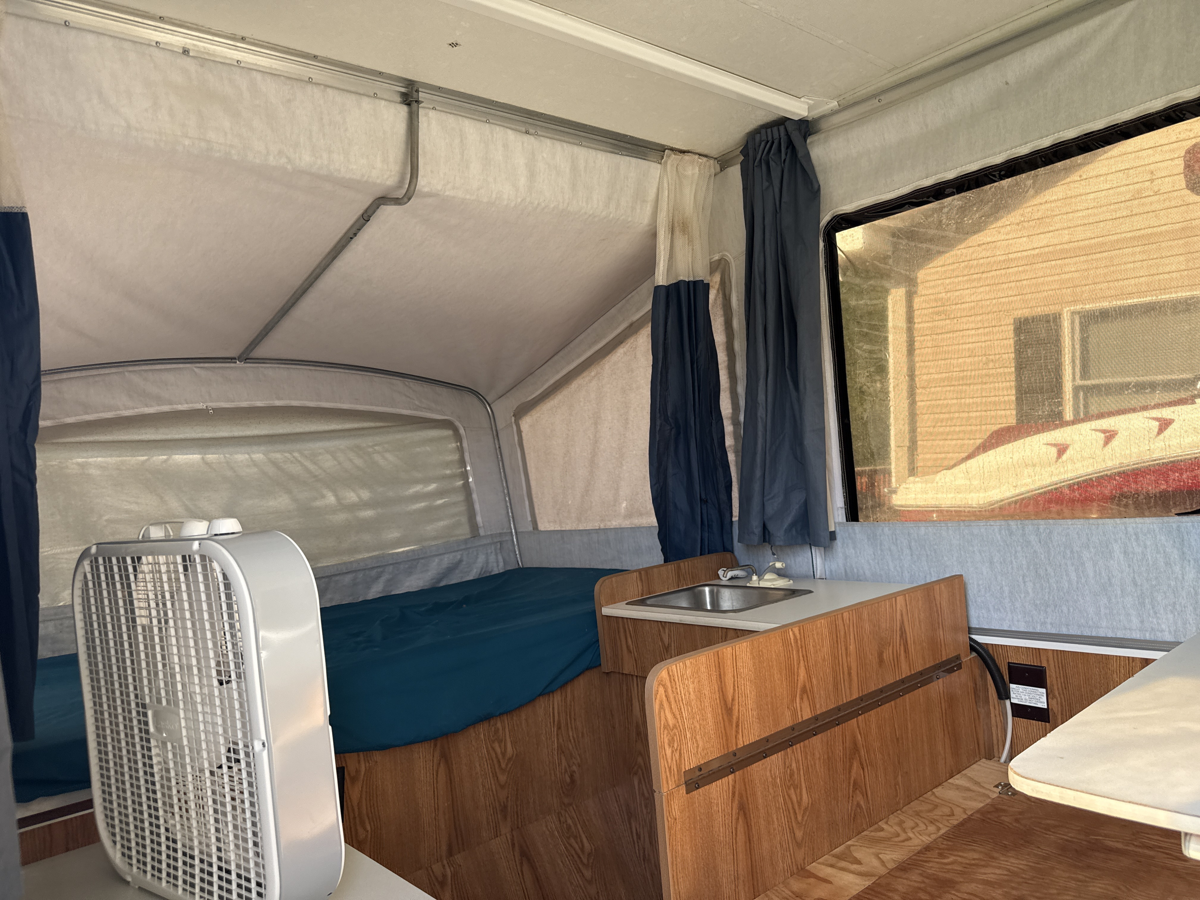 rv rental pop-up camper interior with a bed, small sink, fan, and an outdoor view of a house and boat.. Dutchmen Coleman Lantern 1999