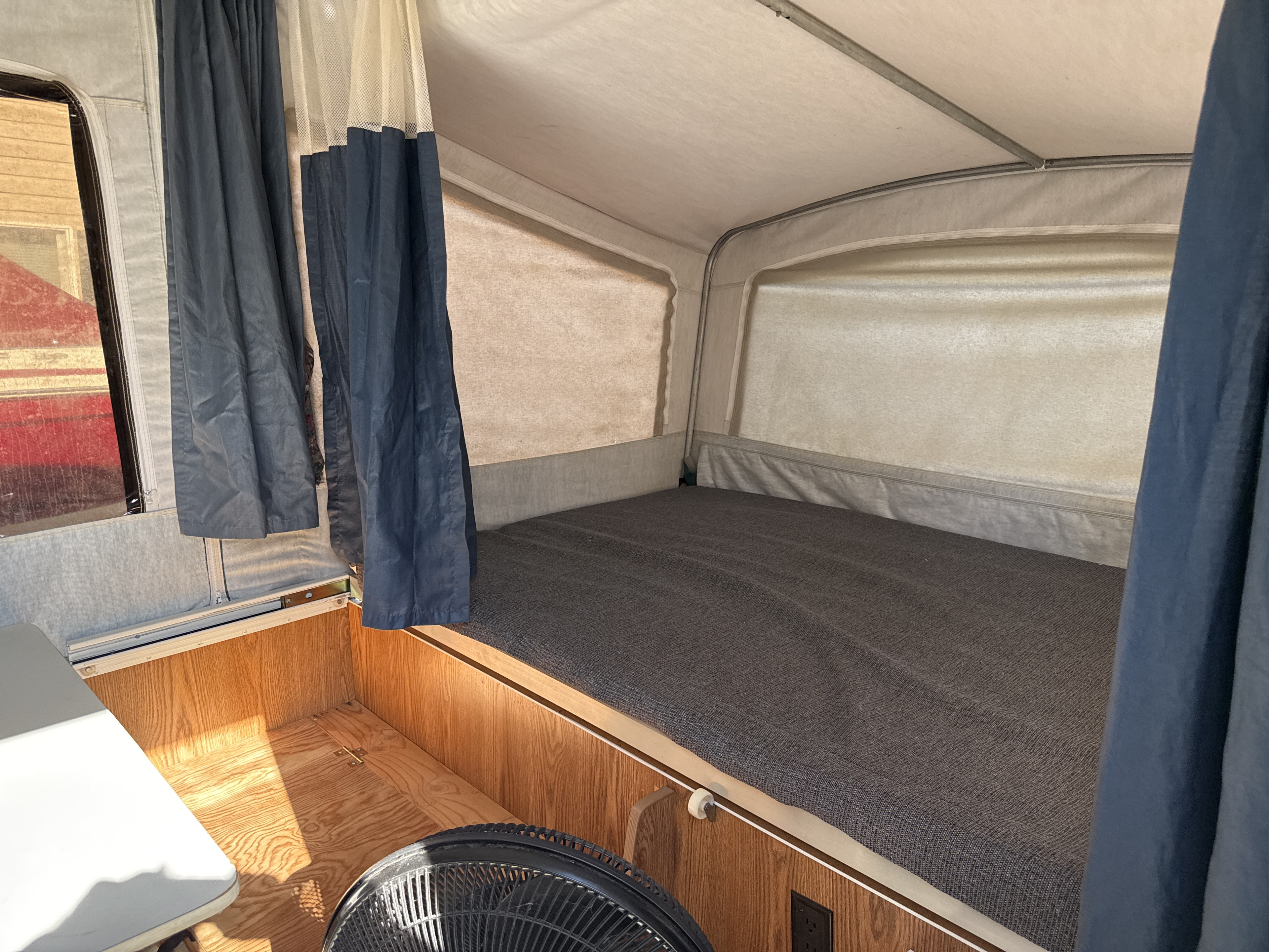 RV rental pop-up camper interior: a bed with gray mattress, blue curtains, and fan, ready for camping.. Dutchmen Coleman Lantern 1999