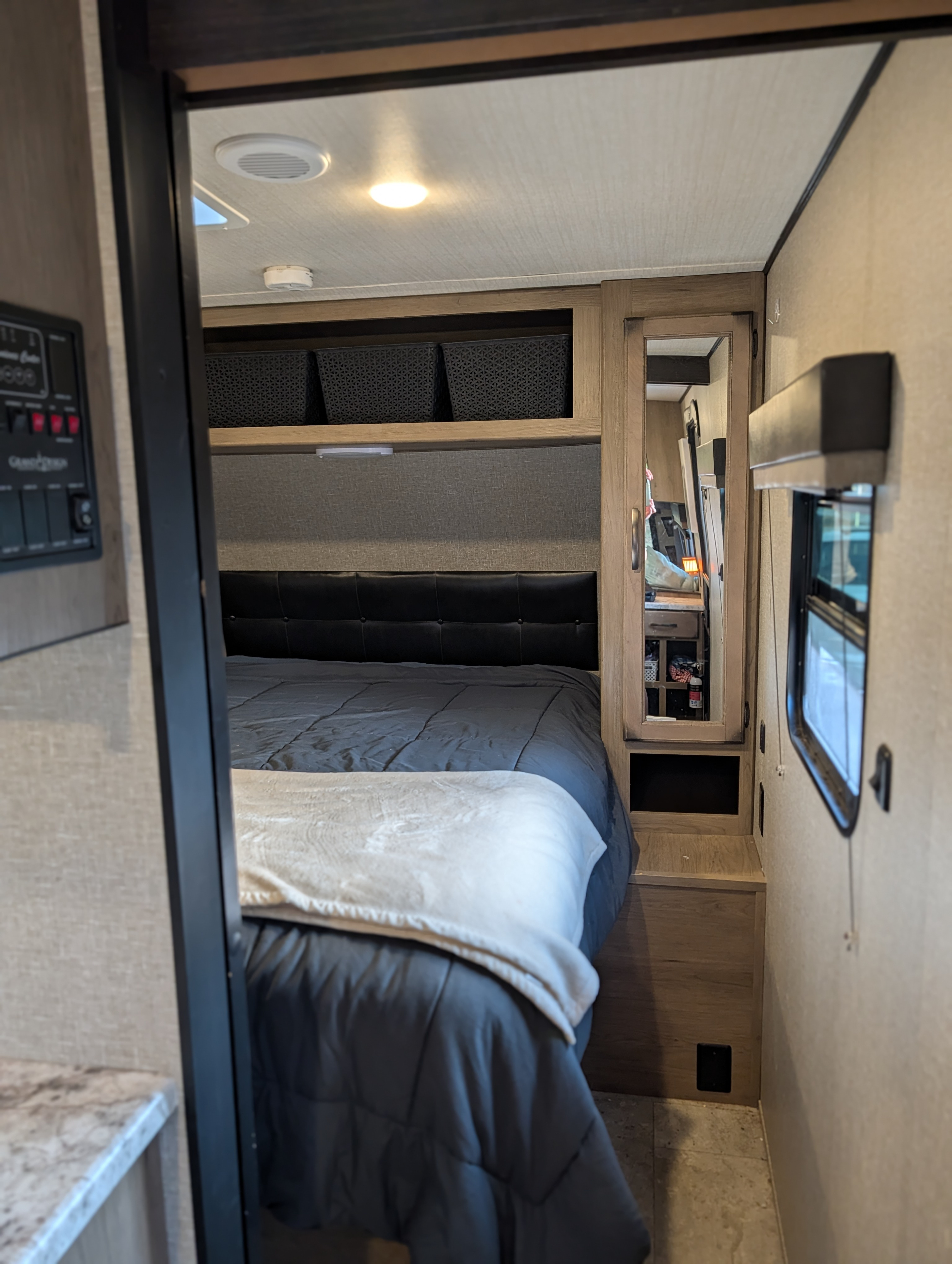 Cozy RV rental bedroom features a comfy bed with a dark comforter, upholstered headboard, overhead storage baskets, and a mirrored cabinet.. Grand Design Transcend 2020