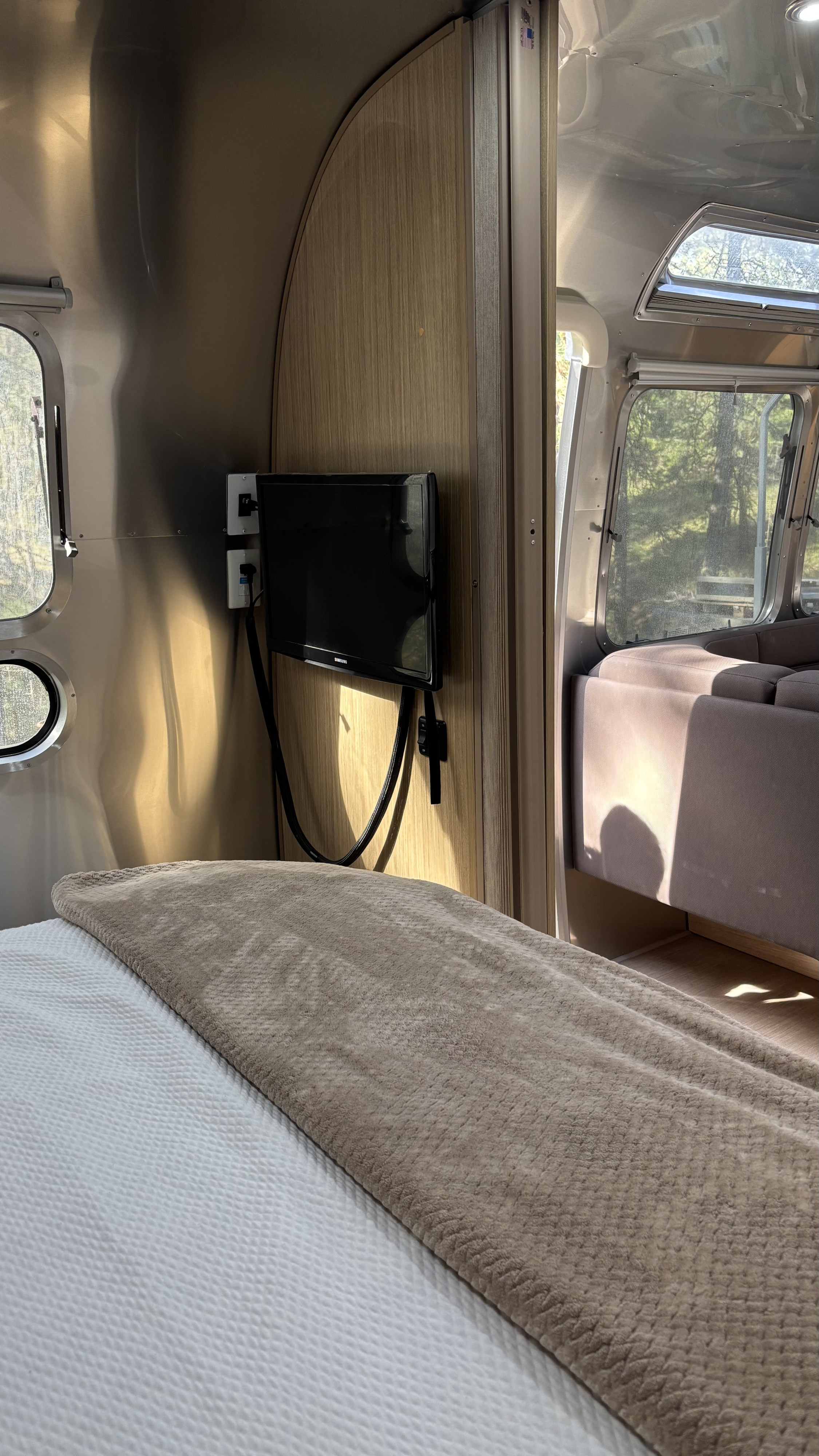 Comfortable bed in an Airstream rv rental bedroom, featuring a TV, metallic walls, and forest views from windows.. Airstream Flying Cloud 2018