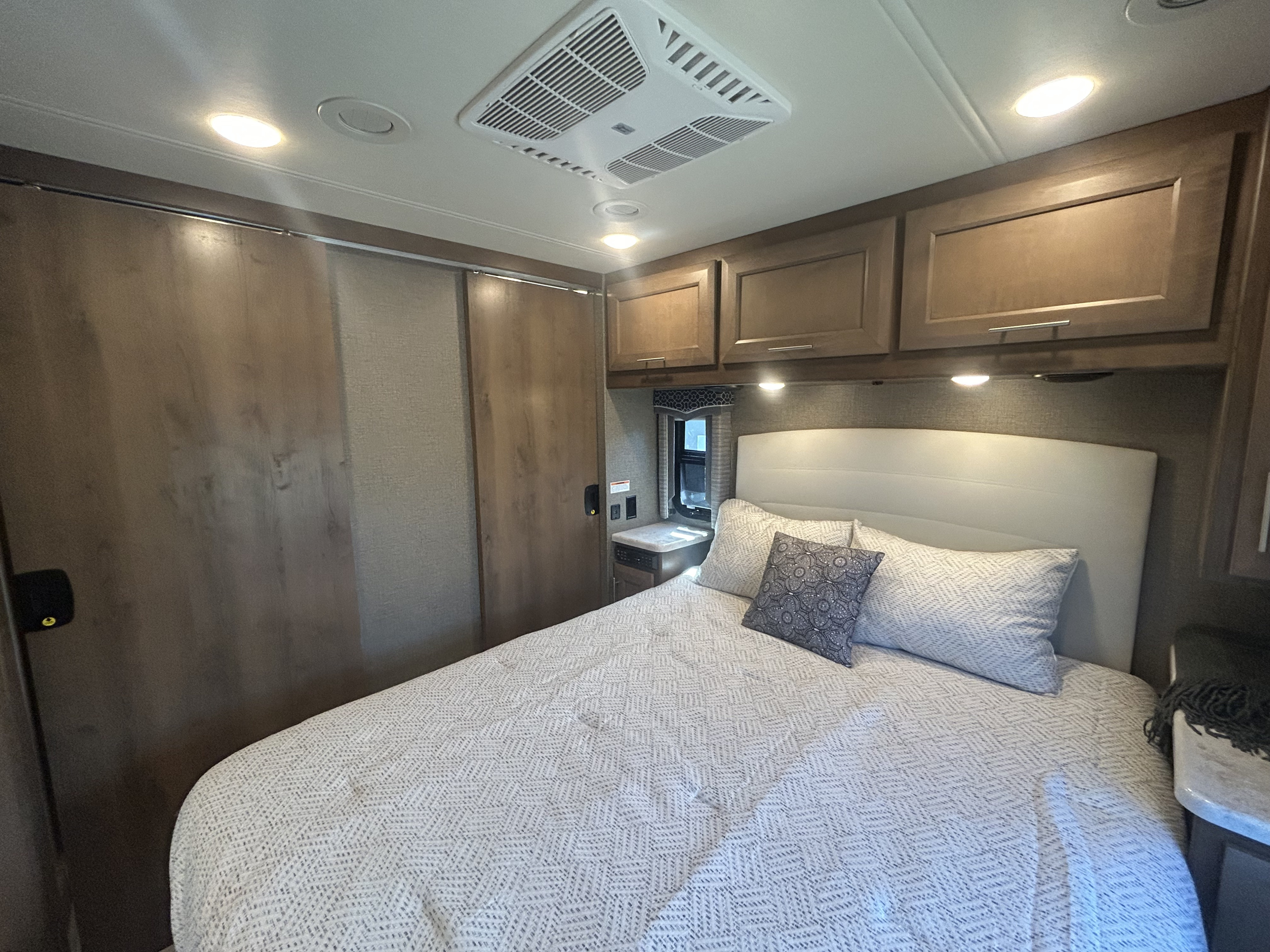 Bright RV rental bedroom featuring a bed with headboard, overhead AC, cabinets, and sliding closet doors.. Thor Magnitude 2021