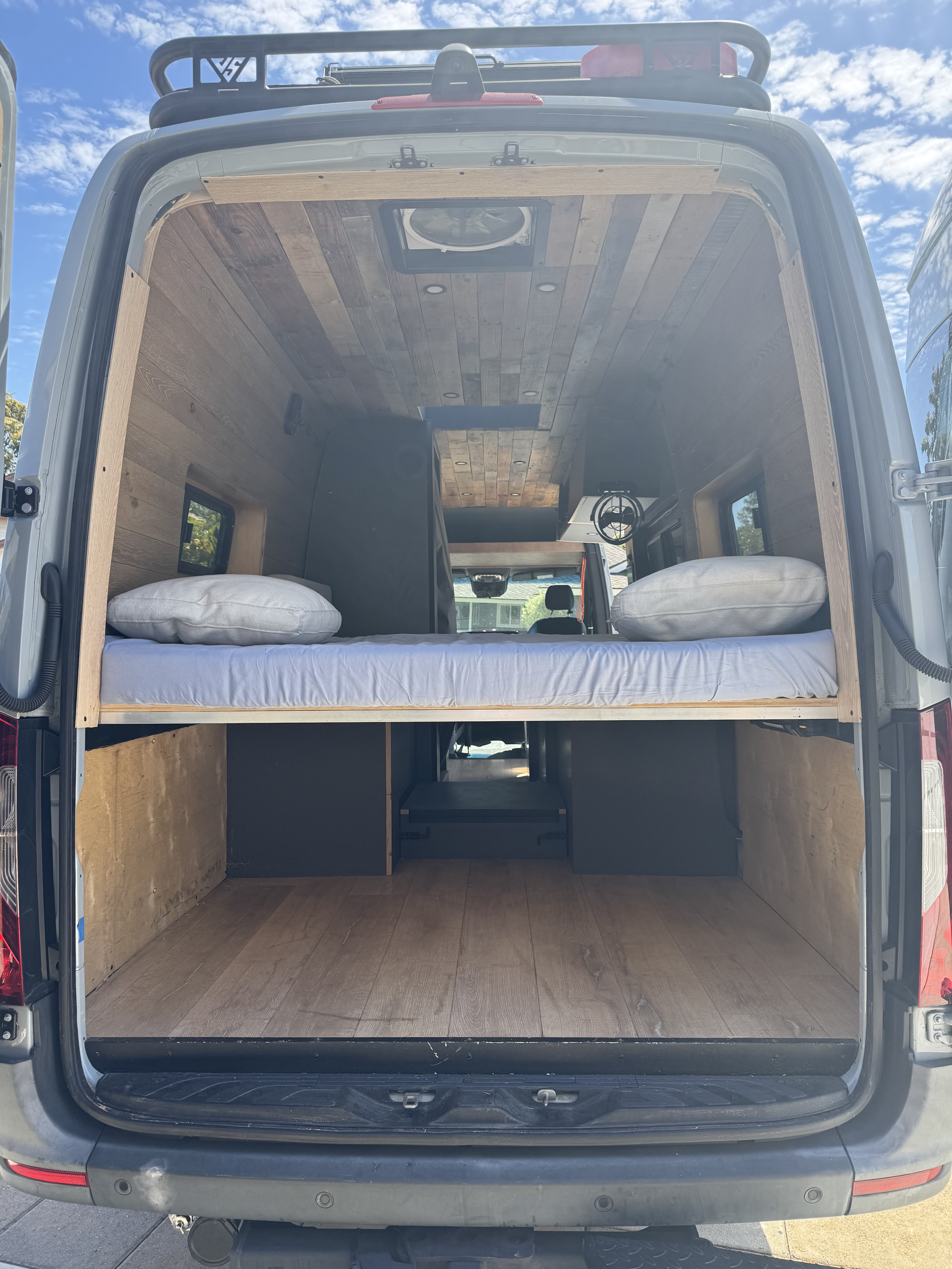 This rv rental reveals a spacious camper van interior with a raised bed, wood-paneled walls/ceiling, and ample storage.. mercedes Mercedes Sprinter 2022