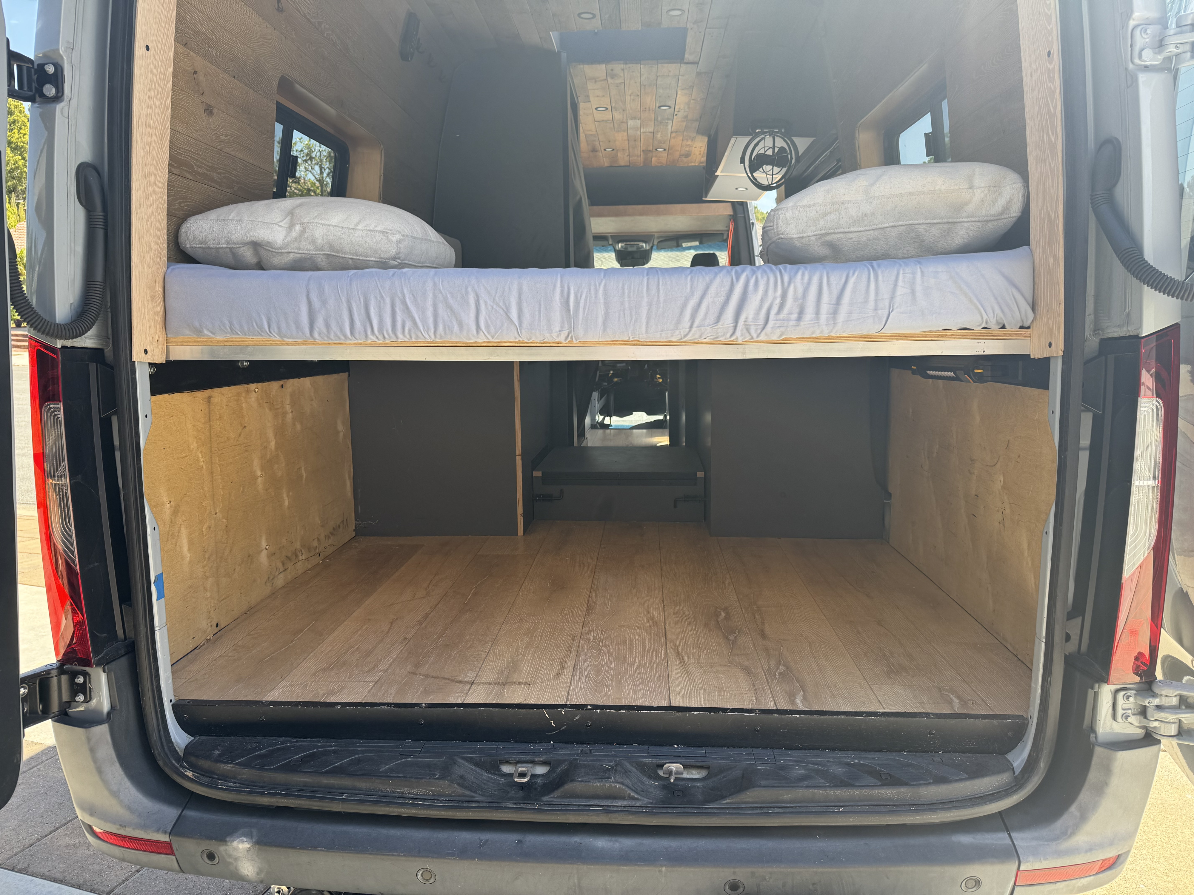 Inside an RV rental: a cozy van conversion with elevated wooden bed, mattress, pillows, and spacious cargo storage.. mercedes Mercedes Sprinter 2022