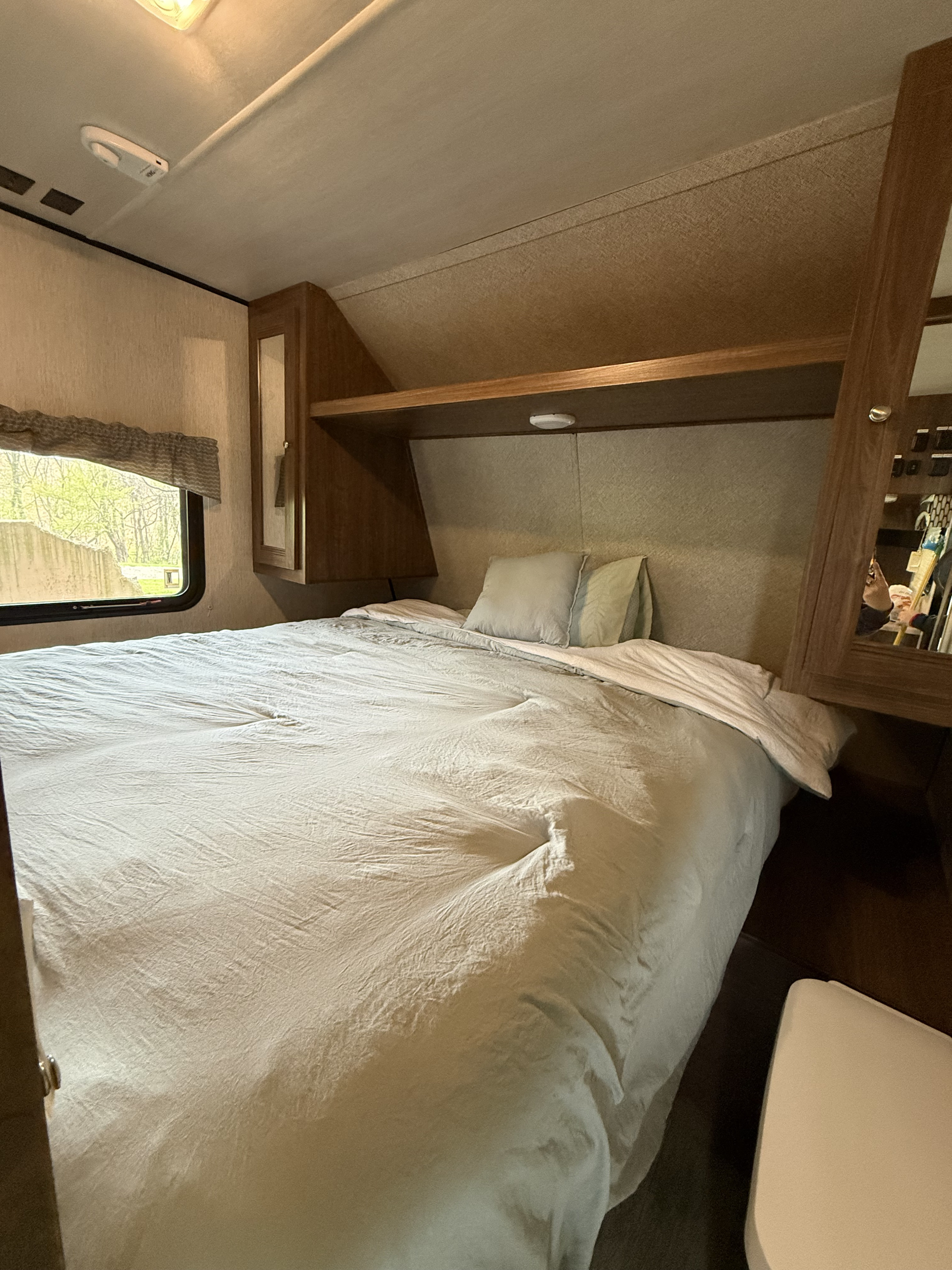 Cozy RV rental bedroom featuring a bed with light bedding, overhead storage, and a window offering a lush green view.. Heartland RVs Prowler Lynx 2018
