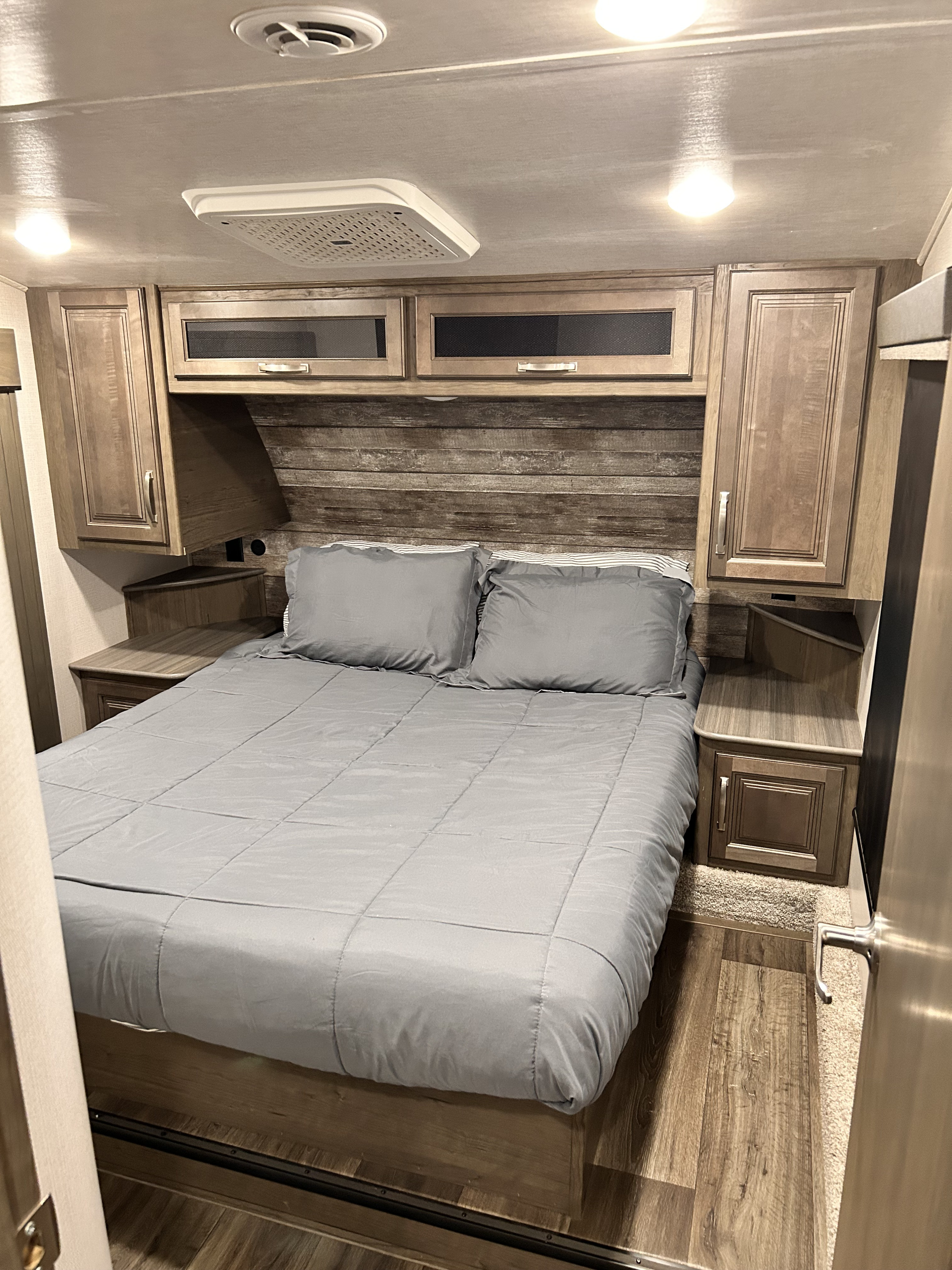 An RV rental bedroom with a comfy gray bed, rustic wood-paneled headboard, overhead storage, and ceiling AC vent.. Keystone RV Cougar 2023