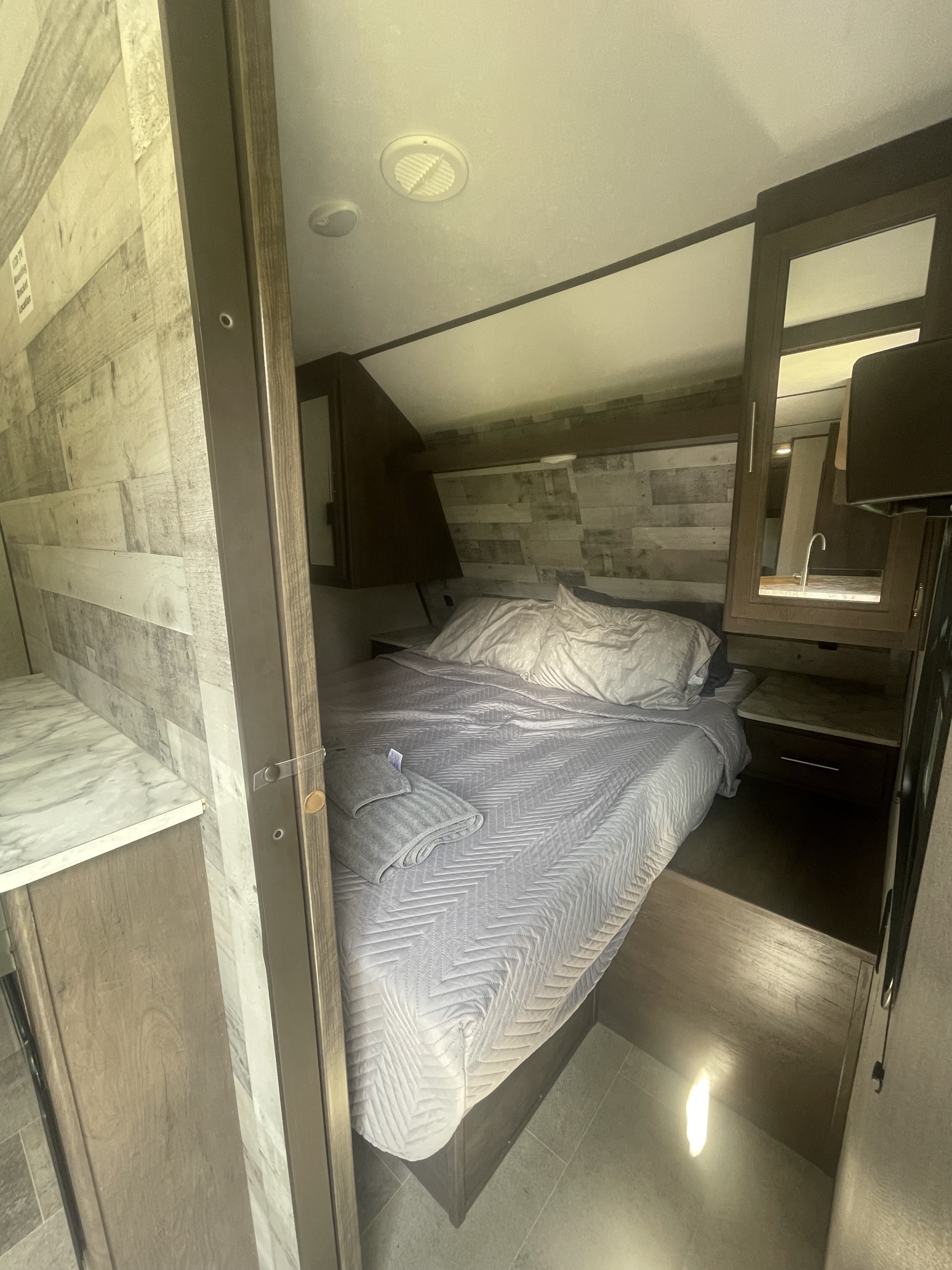 Compact RV rental bedroom with wood-paneled walls, a quilted bed, built-in storage, and a mirrored vanity area.. Dutchmen Coleman Lantern 2021