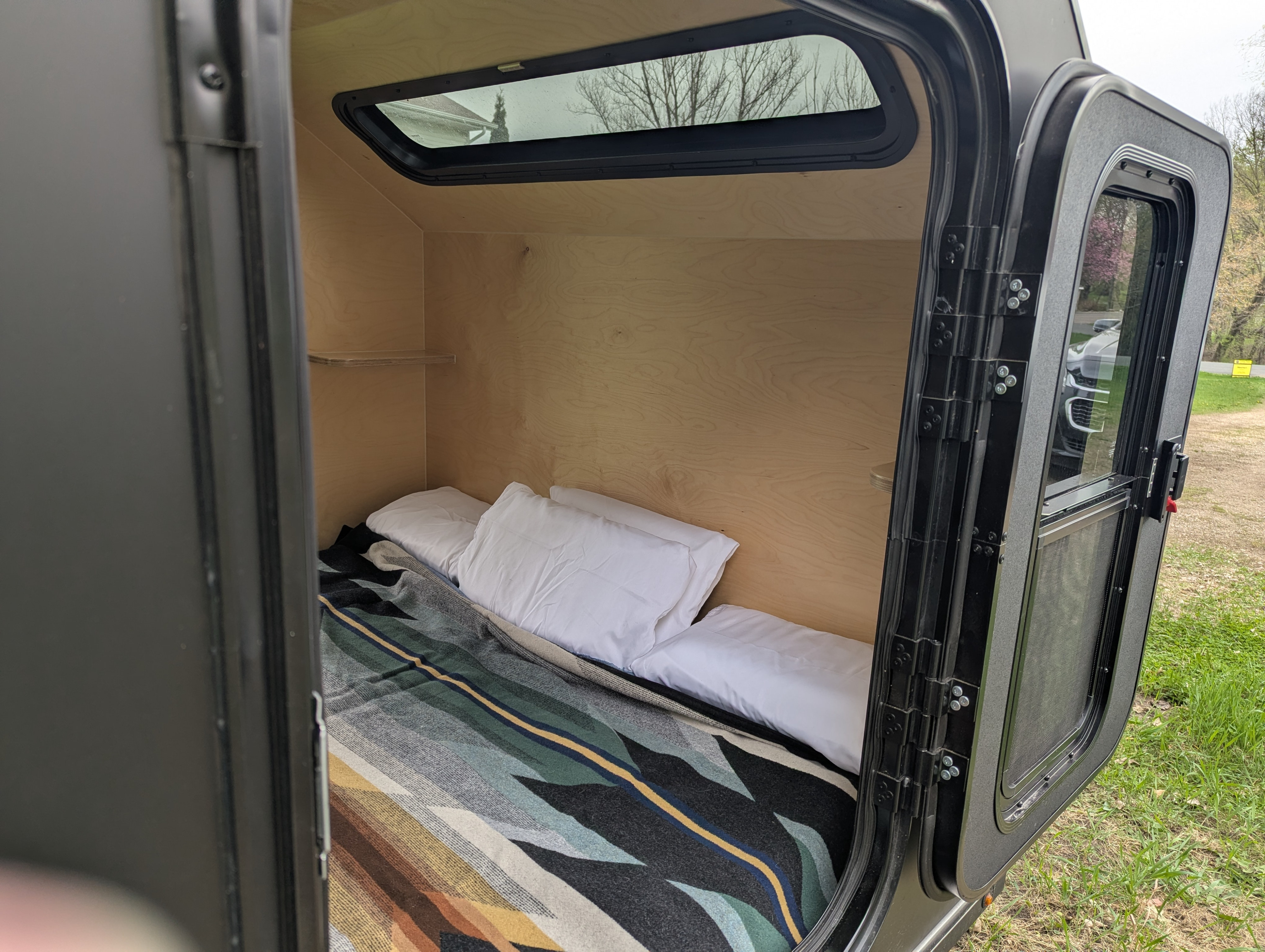 Compact RV rental: A cozy teardrop trailer bedroom with a patterned blanket, white pillows, window, and open door view.. Wander Tears Ranger 2024