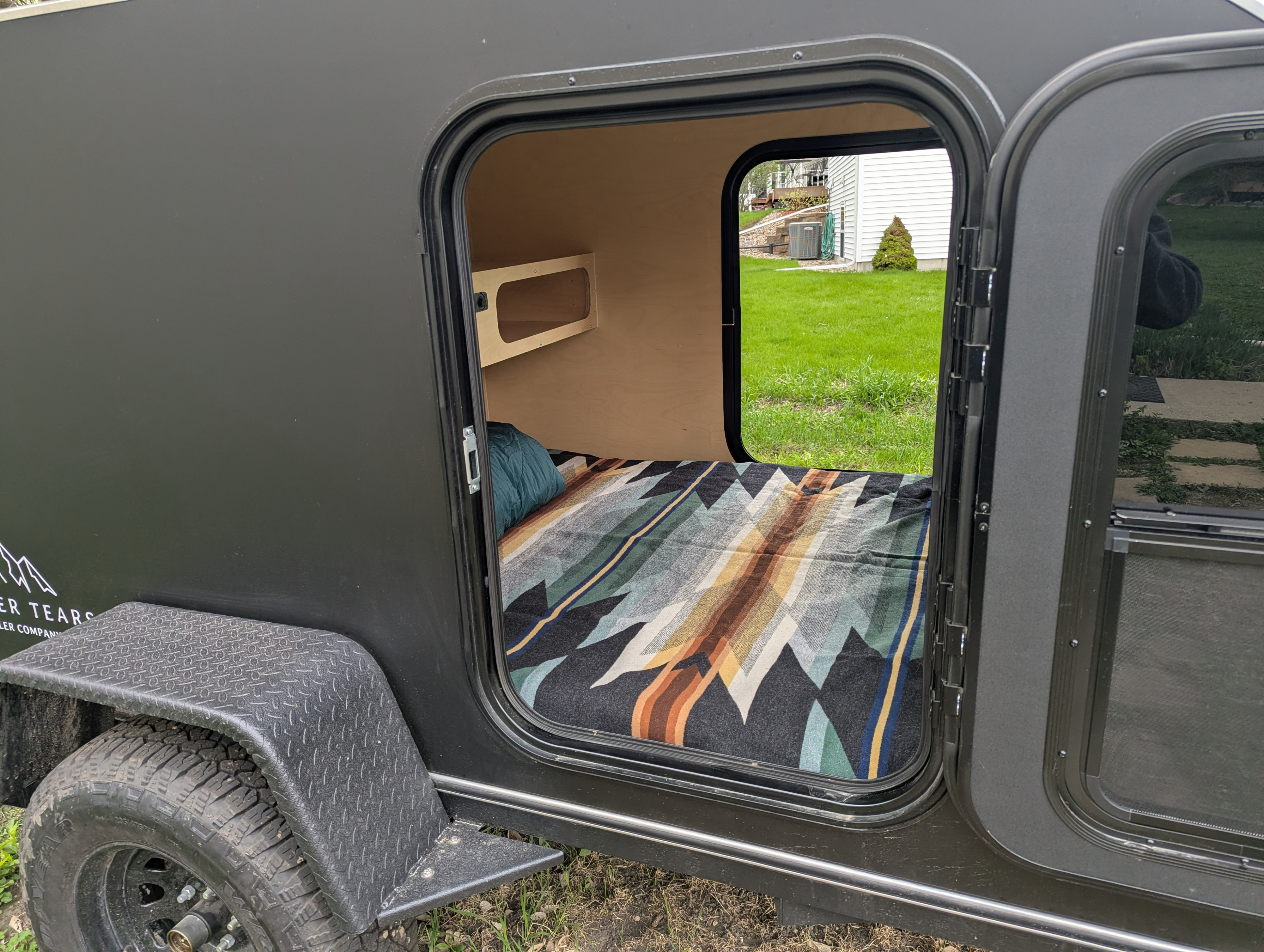 View into a dark grey teardrop camper's open door, showing a cozy bed and shelf. Great for an rv rental adventure.. Wander Tears Ranger 2024