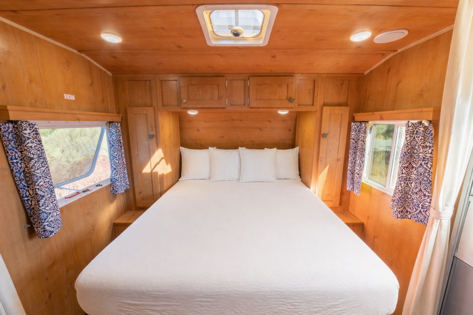 Cozy RV rental bedroom featuring a large bed with white linens, warm wood interior, overhead cabinets, and patterned window curtains.. Other Other 2021