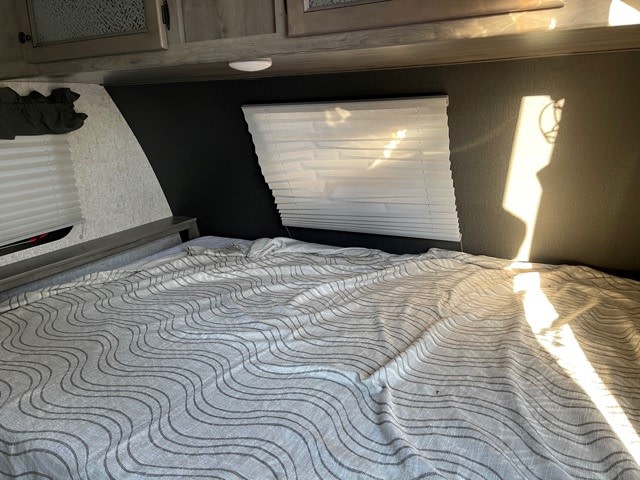 RV rental bedroom with a comfy patterned bed, closed blinds, cabinets above, and sunbeams creating shadows.. Coachmen Apex Nano 2020