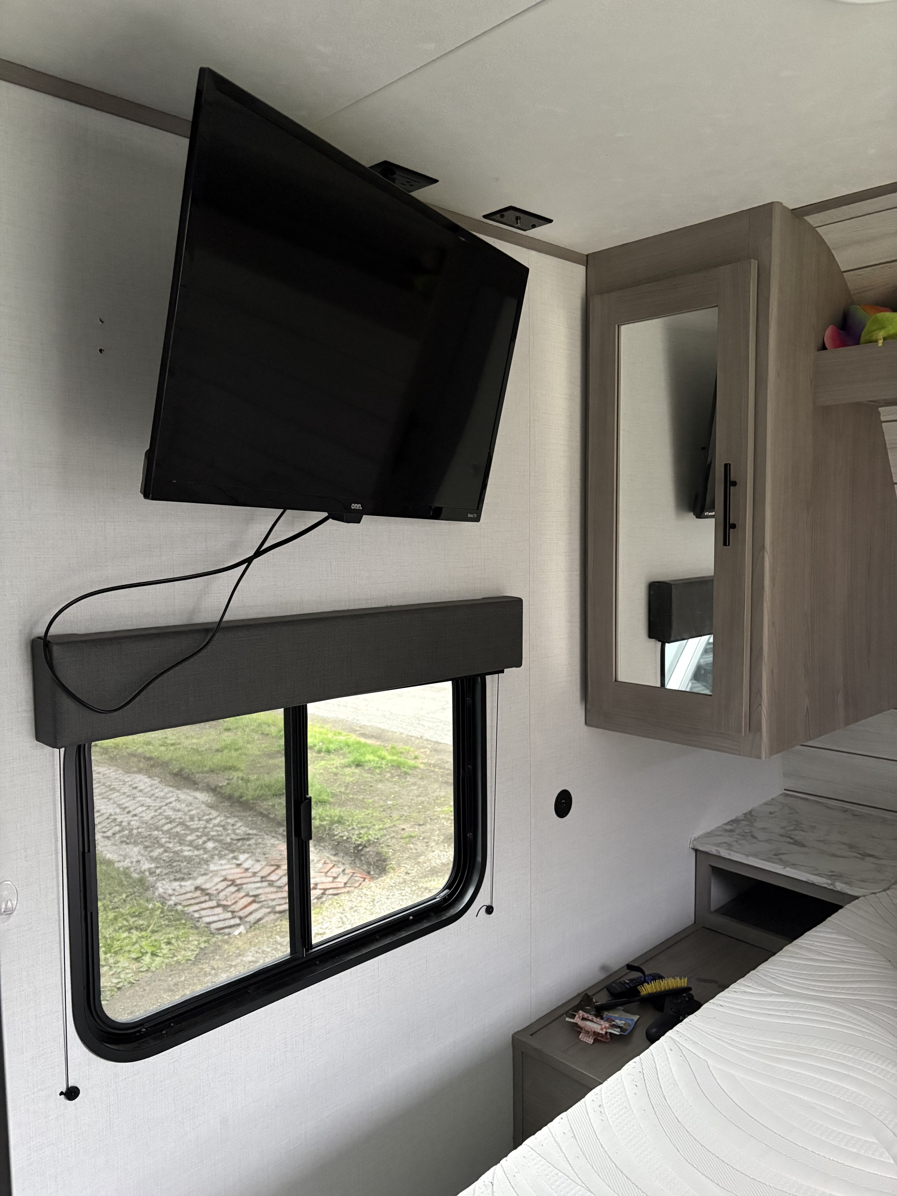 Cozy RV rental bedroom featuring a wall-mounted TV, window with blind, mirrored cabinet, and nightstand area.. Dutchmen Coleman Lantern LT 2024