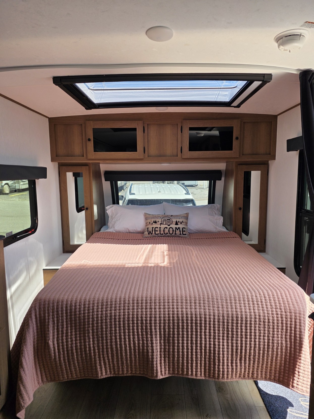 Inside an RV rental, this bright bedroom offers a large bed with a pink quilted spread, "WELCOME" pillow, cabinets, and skylight.. Forest River Coachman Apex Nano 208BHS 2026