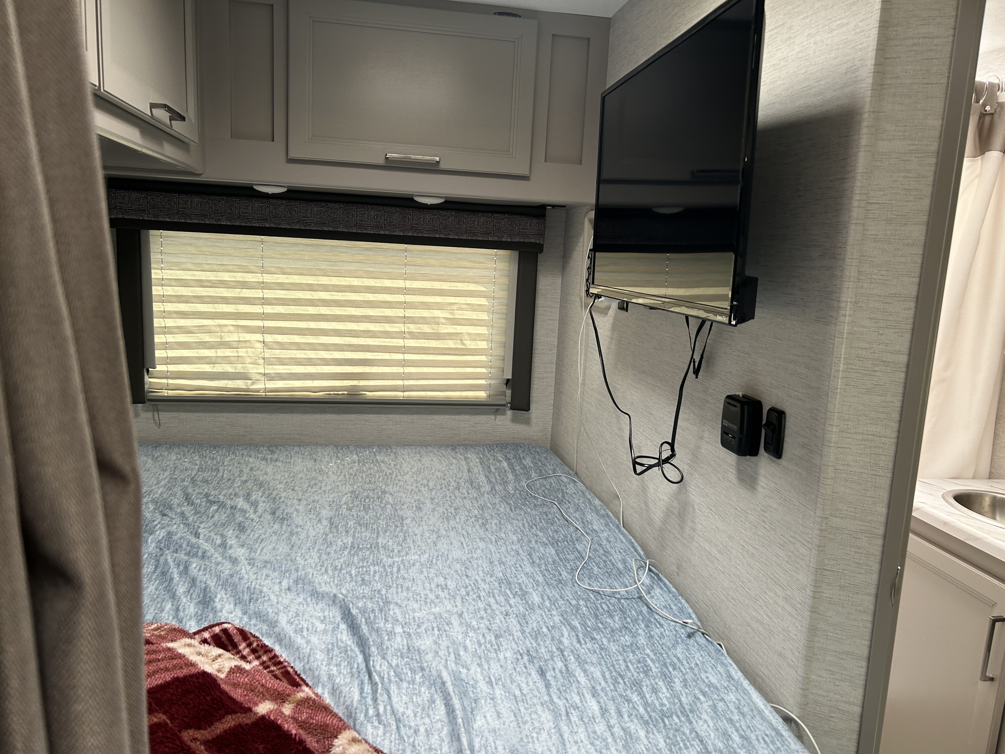 View inside an RV rental showing a compact bedroom with a bed, window, TV, and storage cabinets.. Thor Chateau 2025