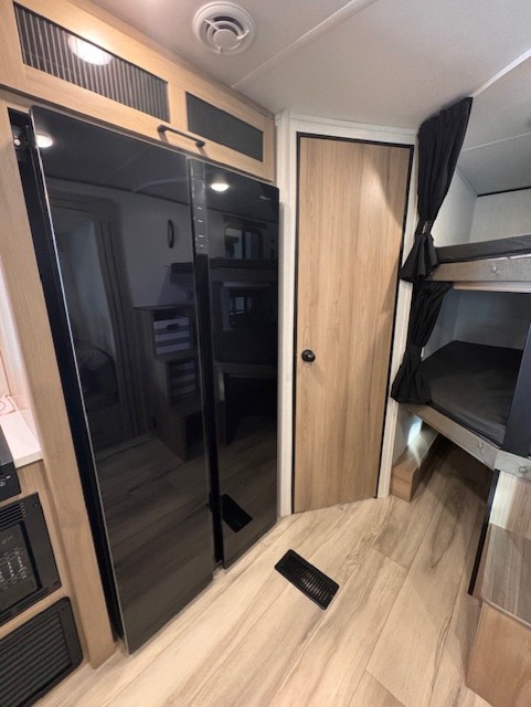 An RV rental interior features a tall black refrigerator, a wooden door, and bunk beds with privacy curtains, ideal for compact living.. Keystone RV Coleman Lantern 2025