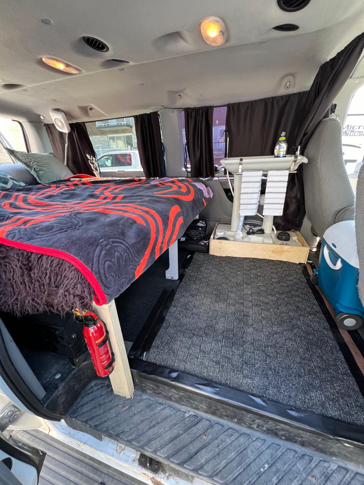 Cozy RV rental camper van interior: bed with red blanket, sink, blue cooler, and fire extinguisher for van life comfort.. Ford Econoline 150 2008
