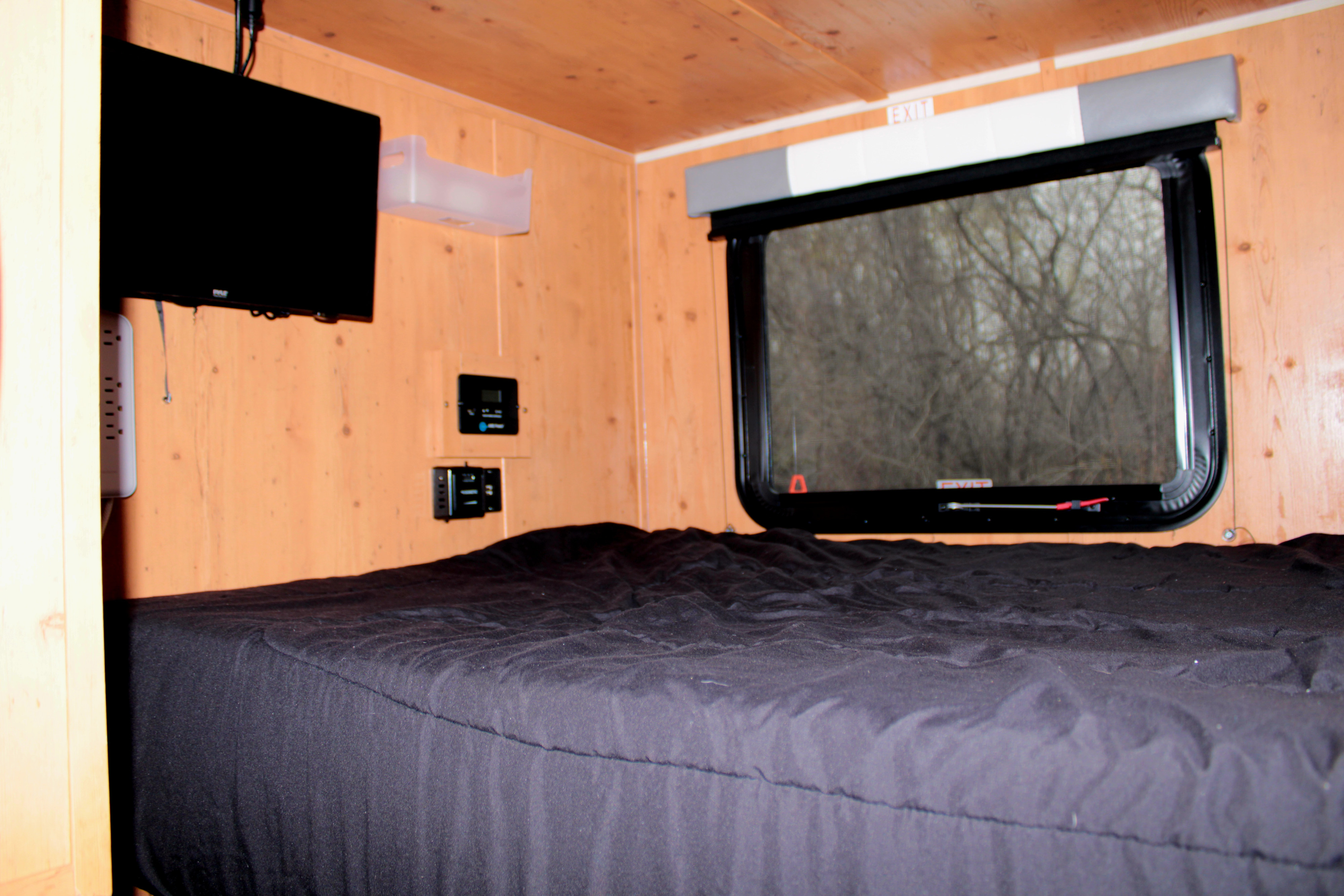 RV rental bedroom: black bed, mounted TV, wood walls, window with blind, controls, & EXIT sign.. Sunset Park RV SunRay 2023