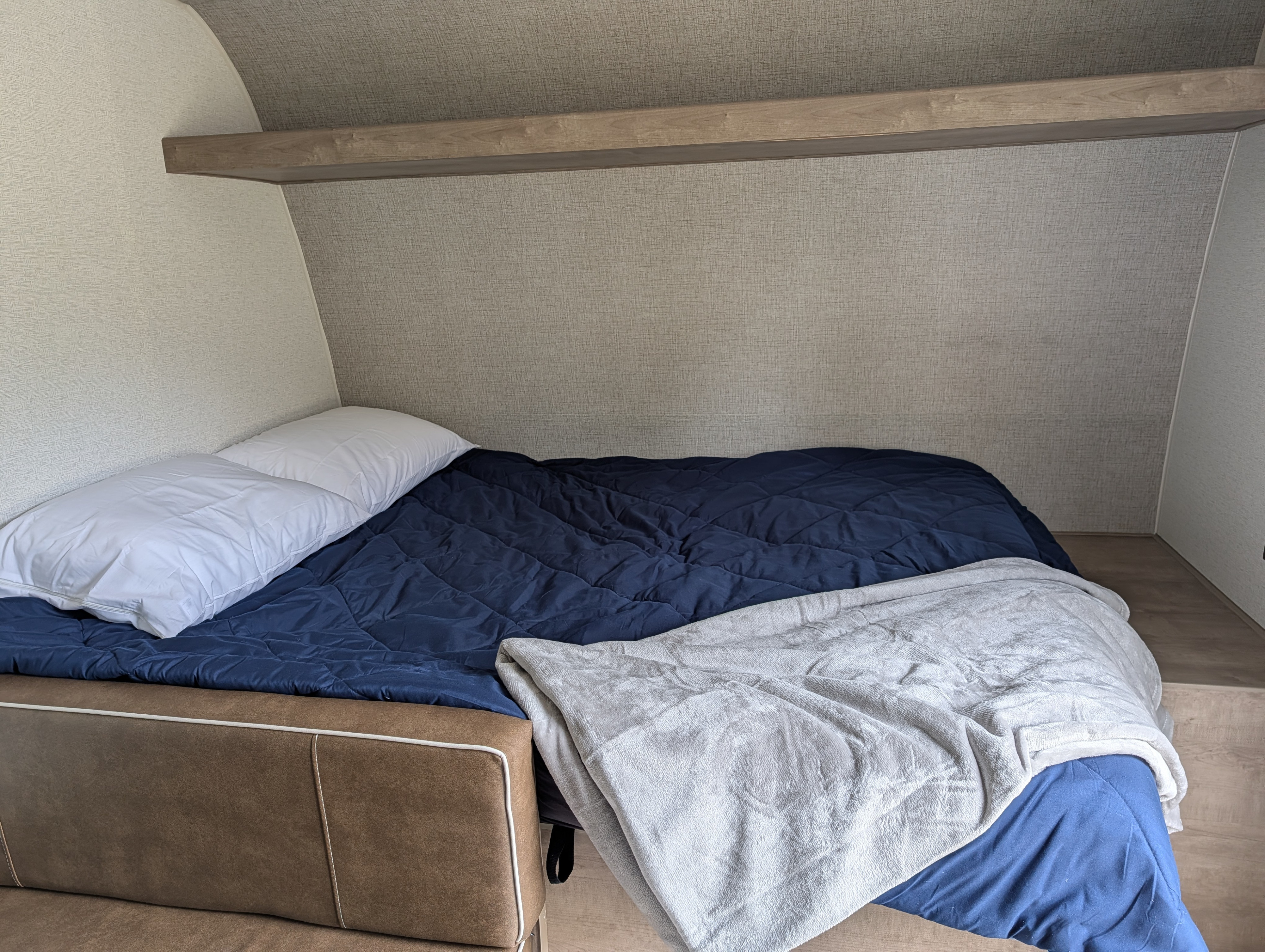 Cozy RV rental bedroom featuring a bed with a dark blue comforter, white pillows, a soft grey blanket, and an overhead shelf.. Keystone RV Springdale Mini 2025