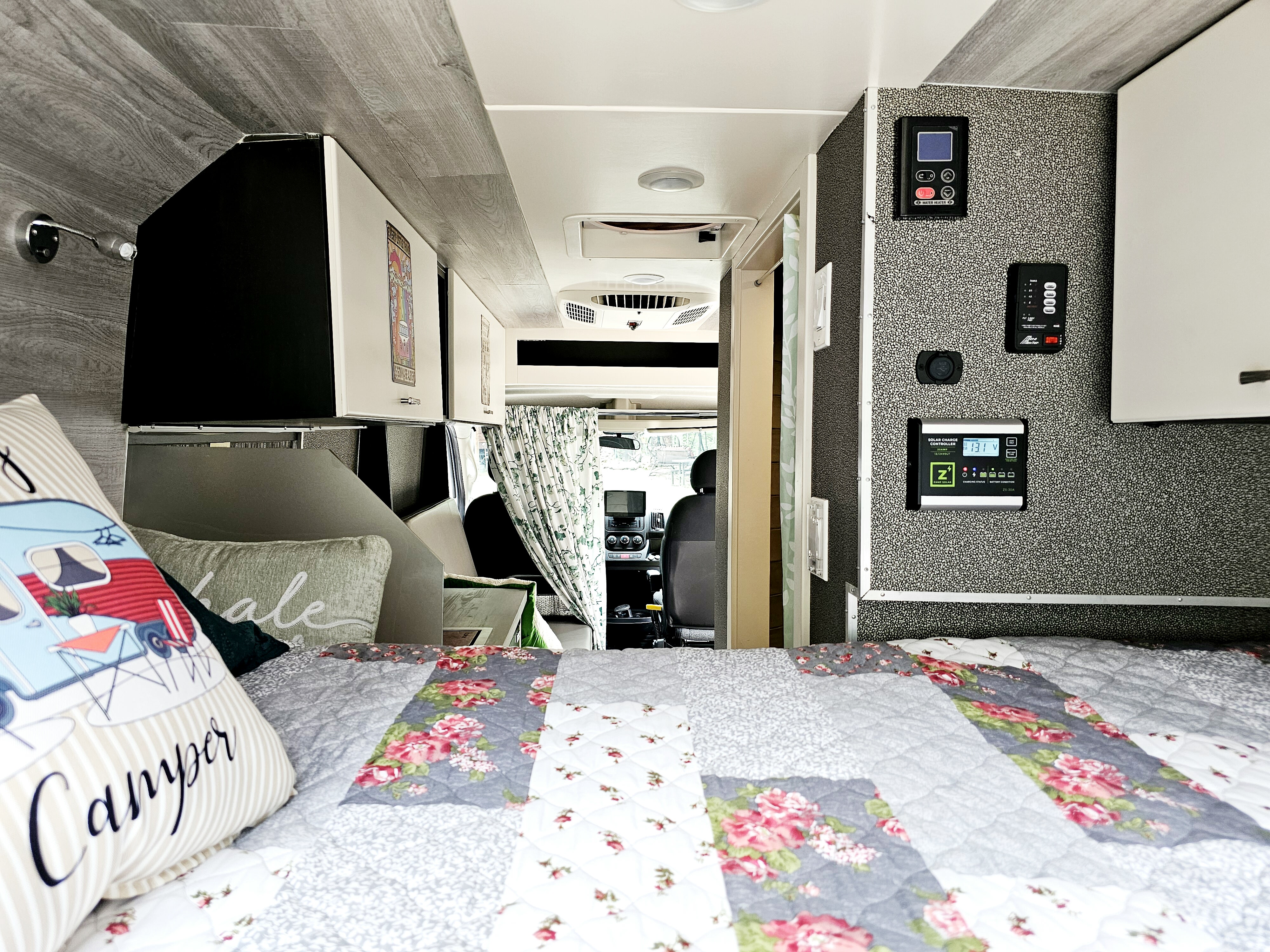 RV rental interior: floral bed, 'Camper' pillow, wall controls, and overhead storage. Driver's cabin visible beyond curtains.. Dodge Ram ProMaster 3500 2018