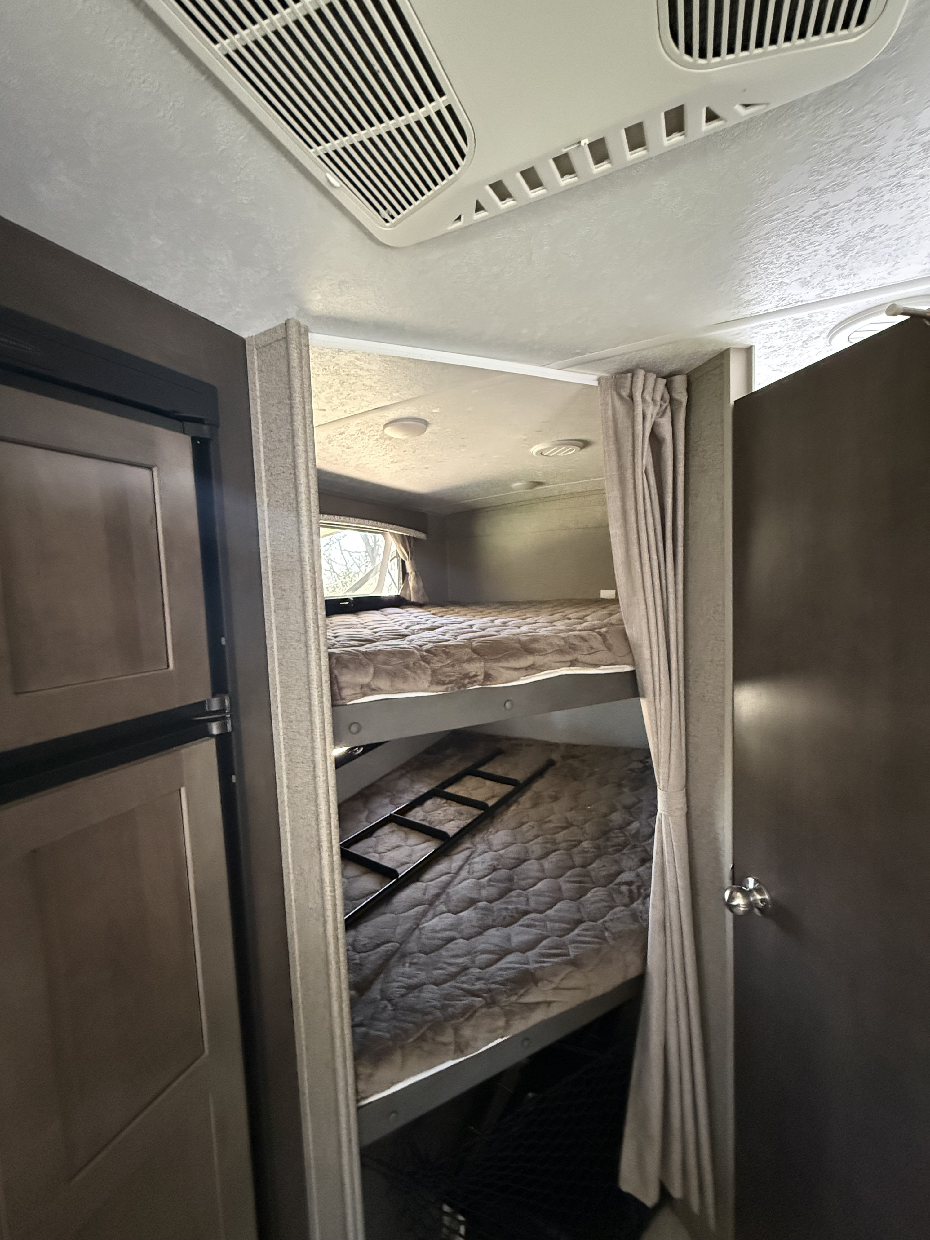 Inside this RV rental, dual bunk beds with a ladder, privacy curtains, and an overhead AC unit offer compact comfort. A fridge is seen left.. Forest River Rockwood Mini Lite 2019