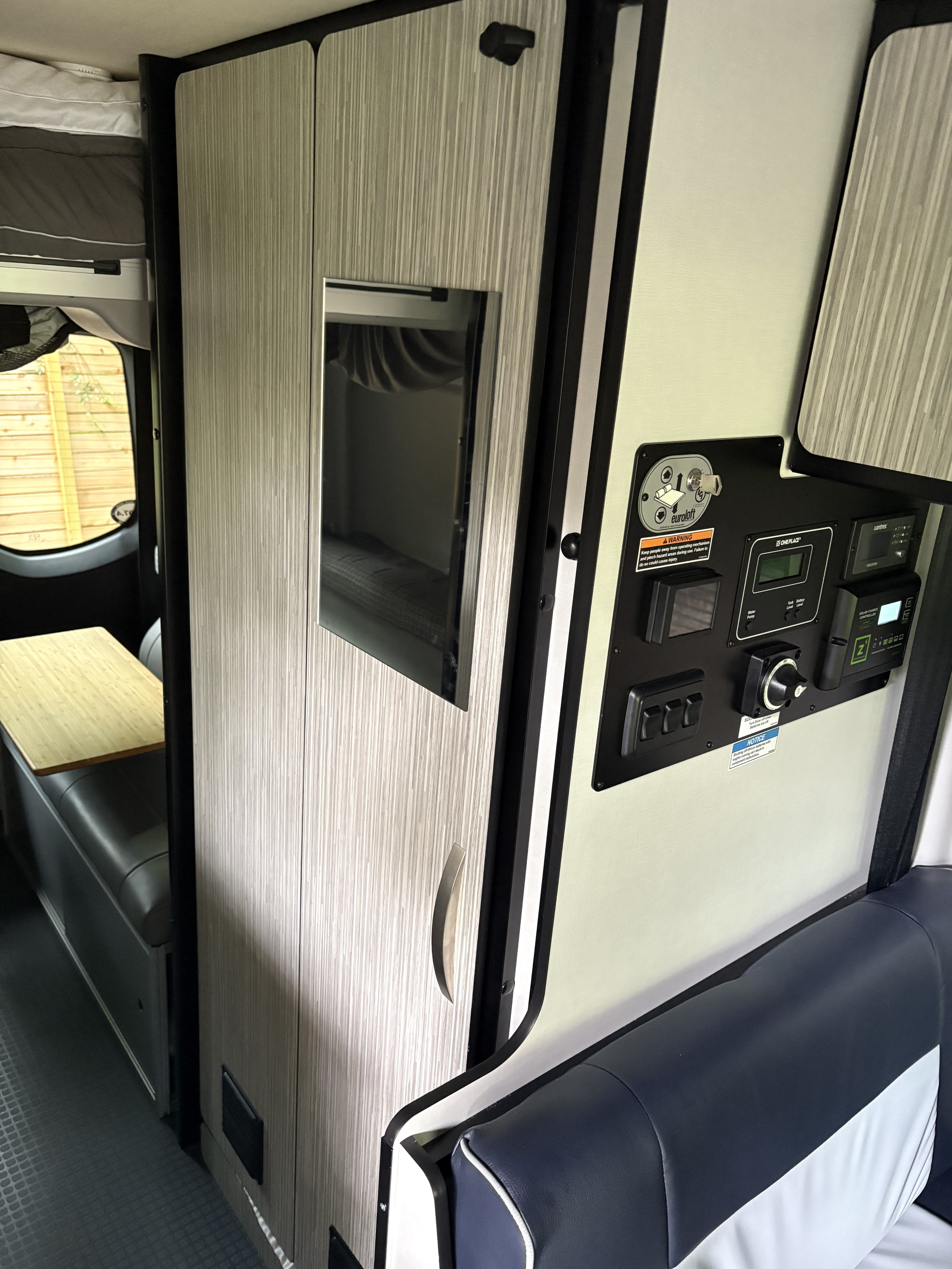 RV rental interior: Control panel for 'eurololft' bed, power, and tank levels, next to dinette & seating, highlighting camper tech.. Winnebago Revel 2024