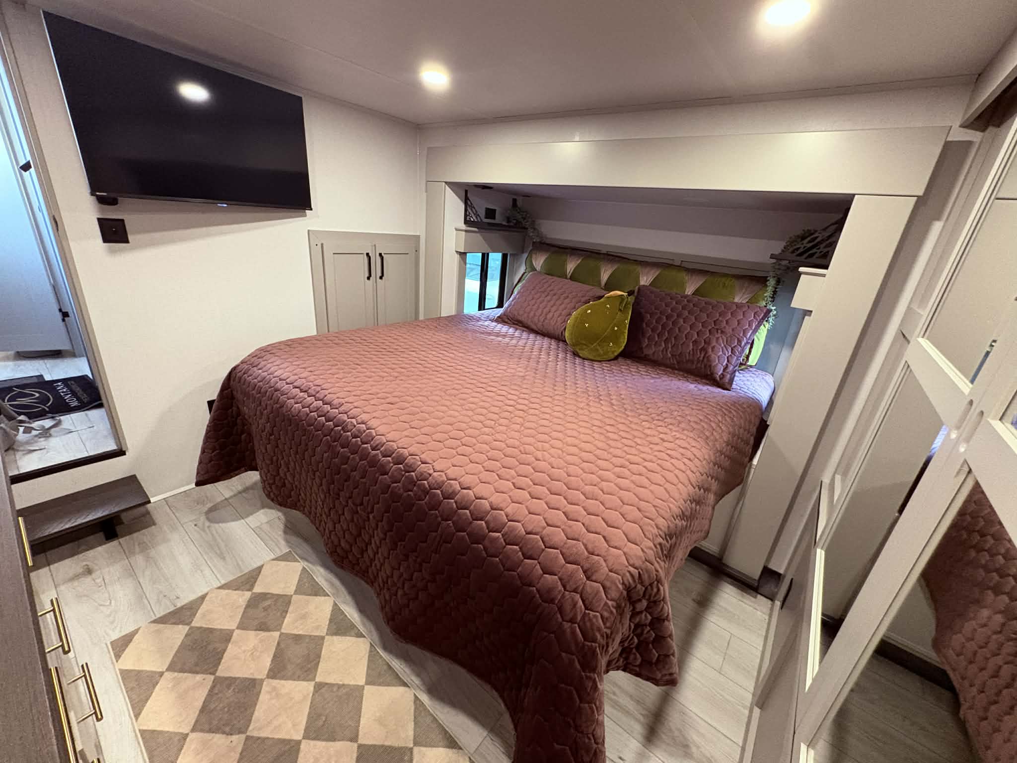This inviting RV rental bedroom features a plush bed with a quilted mauve spread, a mounted TV, and modern built-in storage.. Keystone Montana High Country 2026