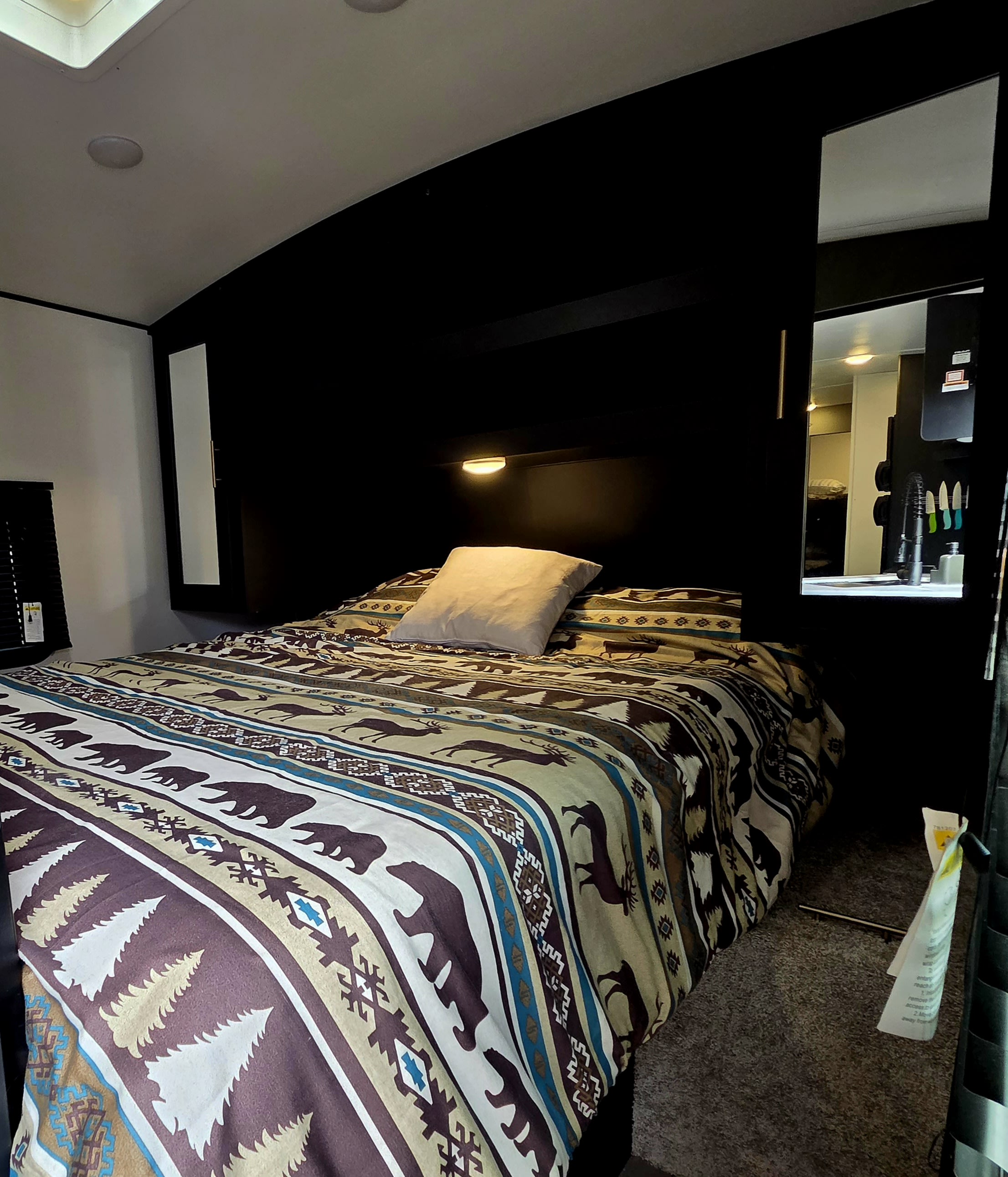This RV rental bedroom features a cozy bed with a rustic bear and deer patterned blanket and a mirror reflecting a kitchen.. Keystone RV Springdale 2022