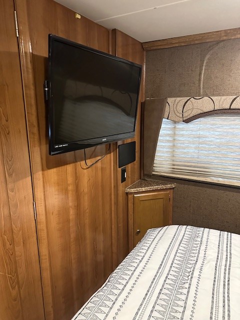 This RV rental bedroom has a mounted TV on a wood wall, a window with blinds, and a bed with a striped comforter.. 1GB6GUCG1H1118350 Leprechaun 2017