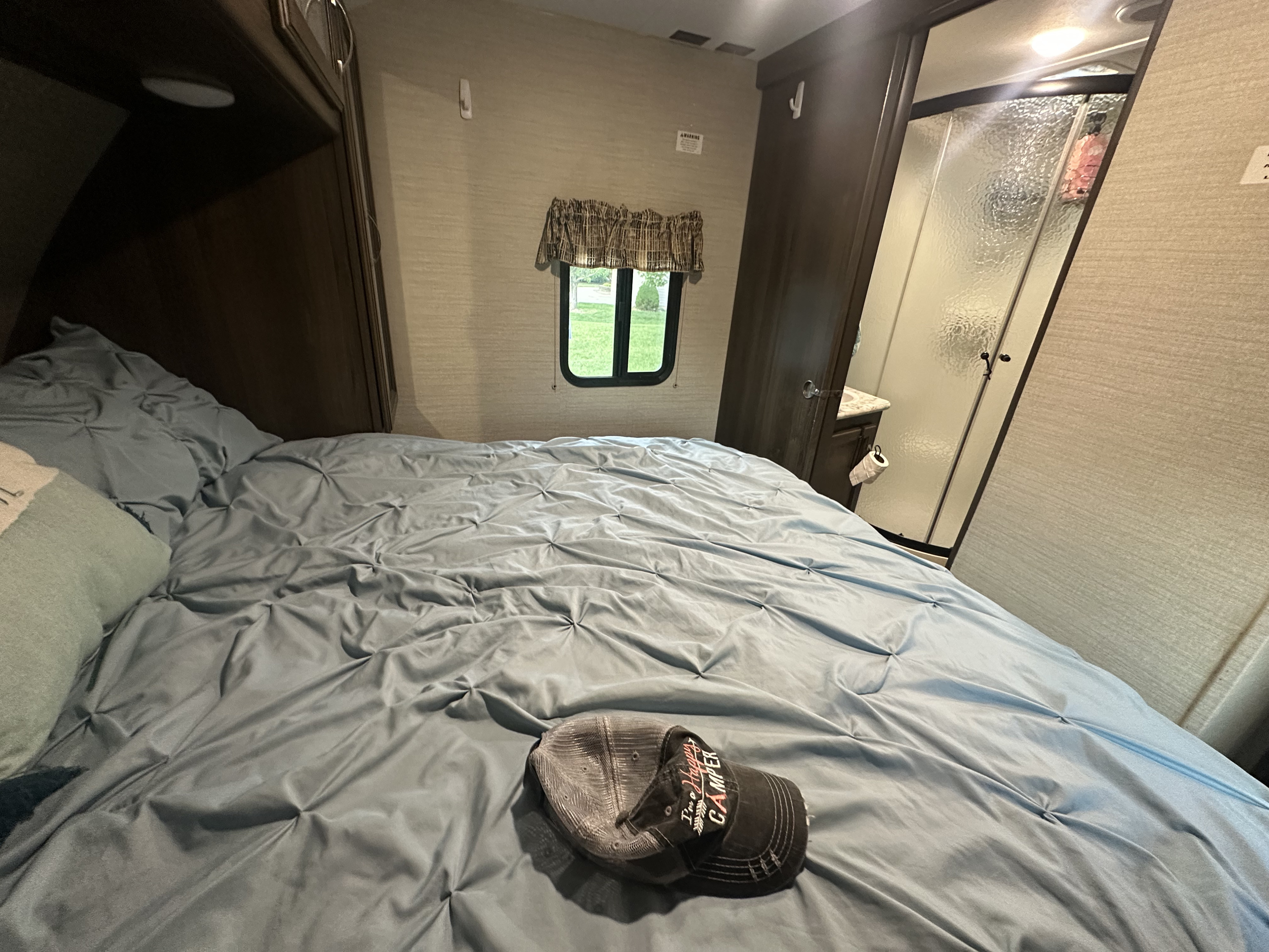 Cozy RV rental bedroom with a blue bedspread, a window showing grass, and a peek at the bathroom. A hat is on the bed.. Keystone RV Bullet Ultra Lite 2019