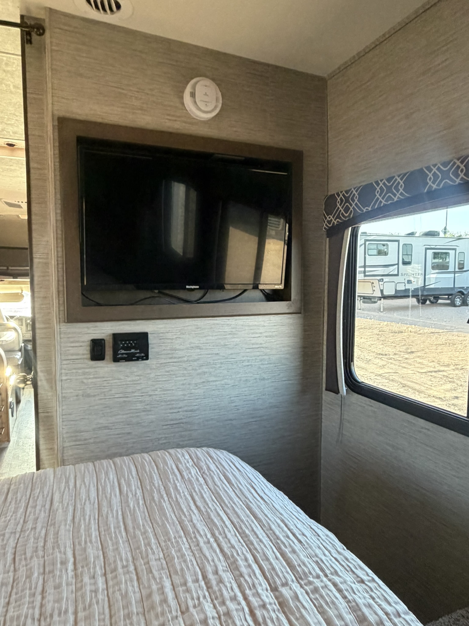 Cozy RV rental bedroom interior with mounted TV, controls, and a window view of another RV. Perfect for your camping trip.. Thor Chateau 2020