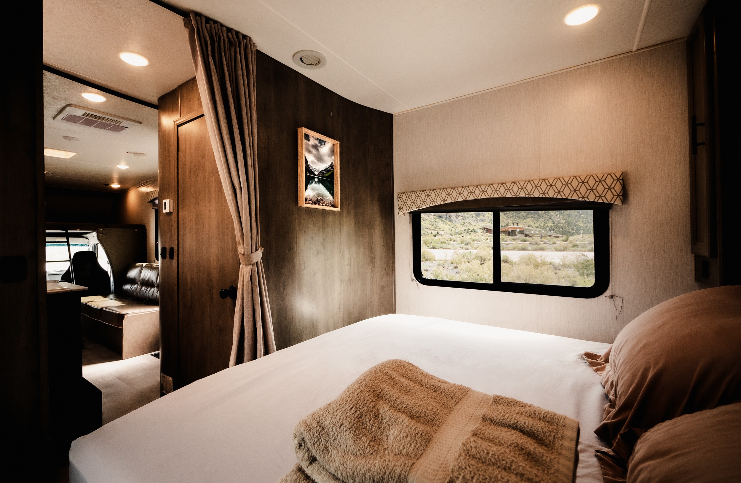 Cozy RV rental bedroom with a white bed, brown pillows, and window overlooking a dry, mountainous landscape. Doorway to living area.. Coach House FREELA 2020
