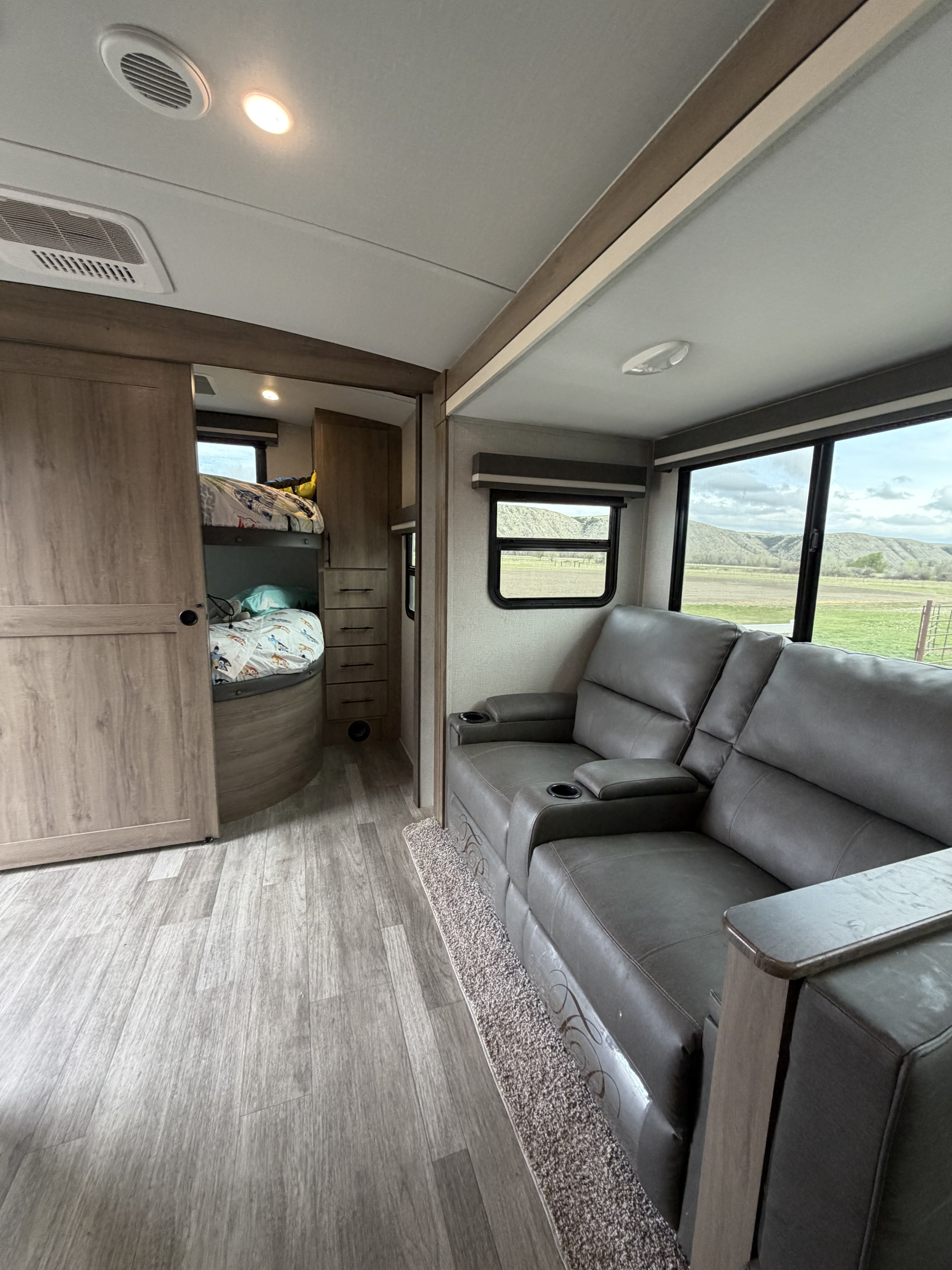 An RV rental's cozy interior, featuring gray recliners, bunk beds, and a scenic window view of distant hills.. Grand Design Imagine 2022