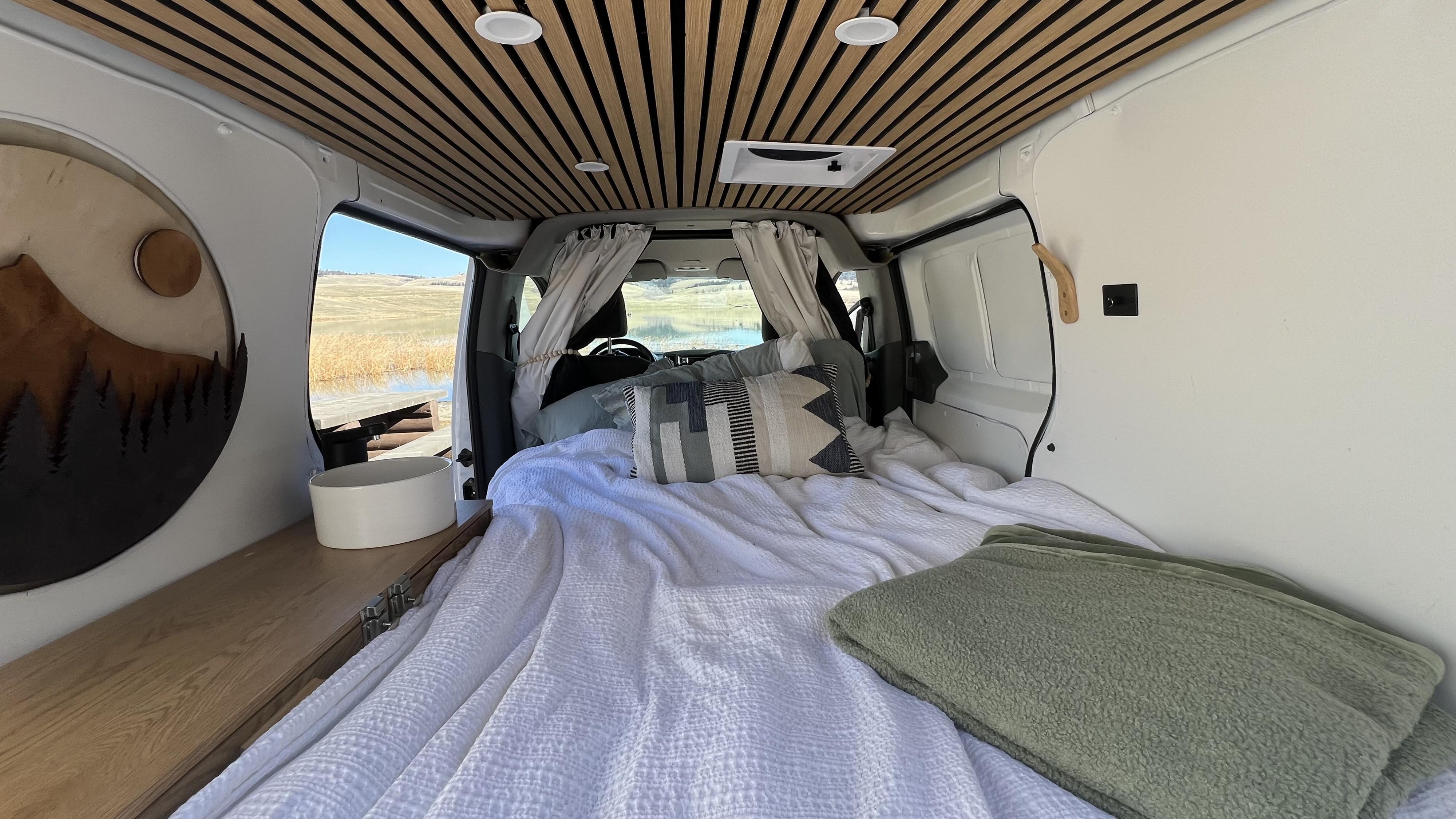Interior of an rv rental: inviting bed, sleek wood-slat ceiling, and scenic lake vista. Road trip ready.. Nissan NV200 2020