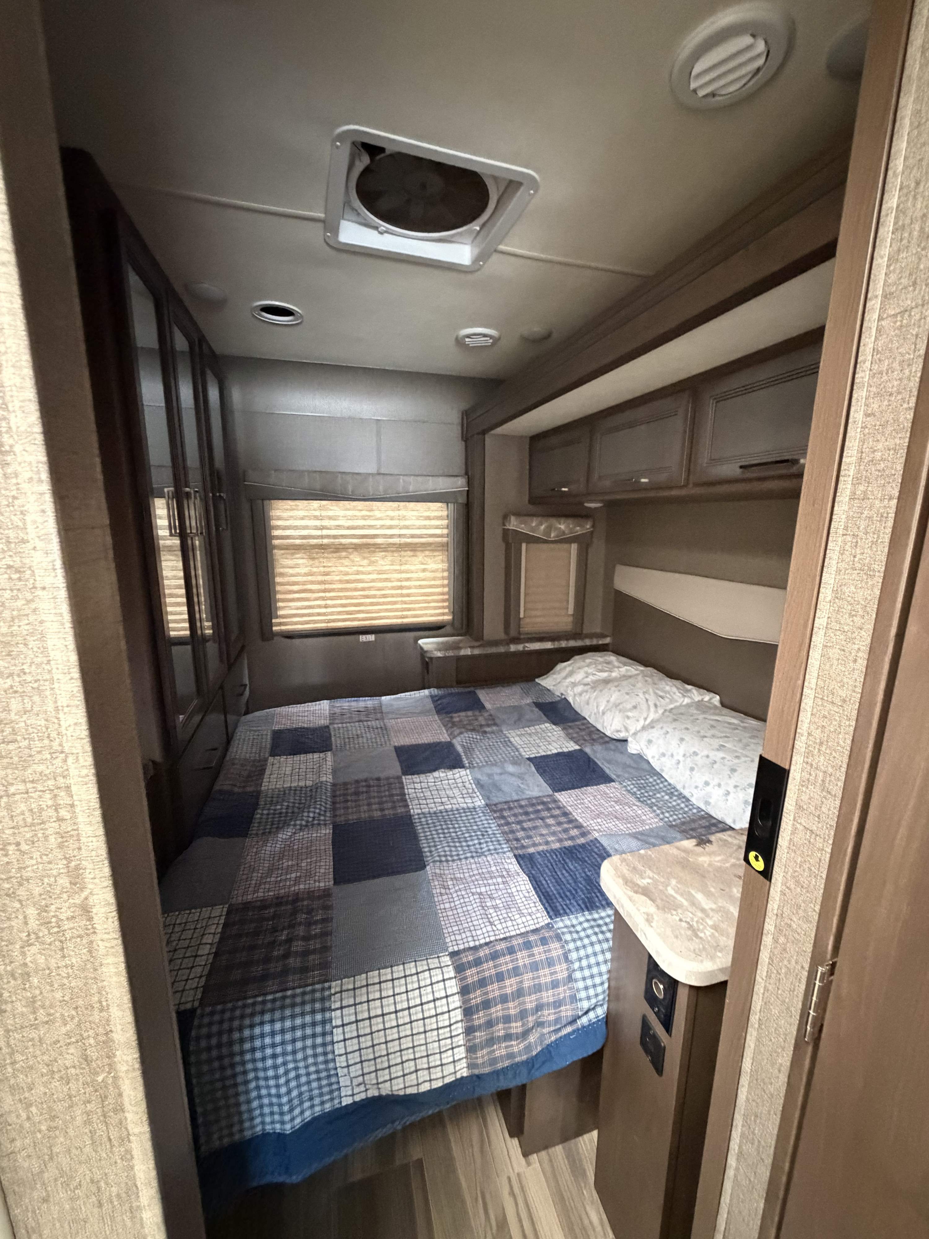 Cozy RV rental bedroom featuring a bed with a patchwork quilt, overhead storage, window blinds, and a ceiling vent.. Thor A.C.E. 2020